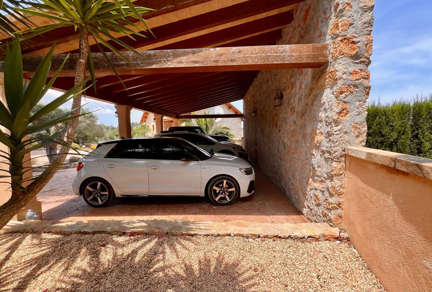 A series of luxury vehicles parked under a desert-style covered carport with stone columns and gravel landscaping.