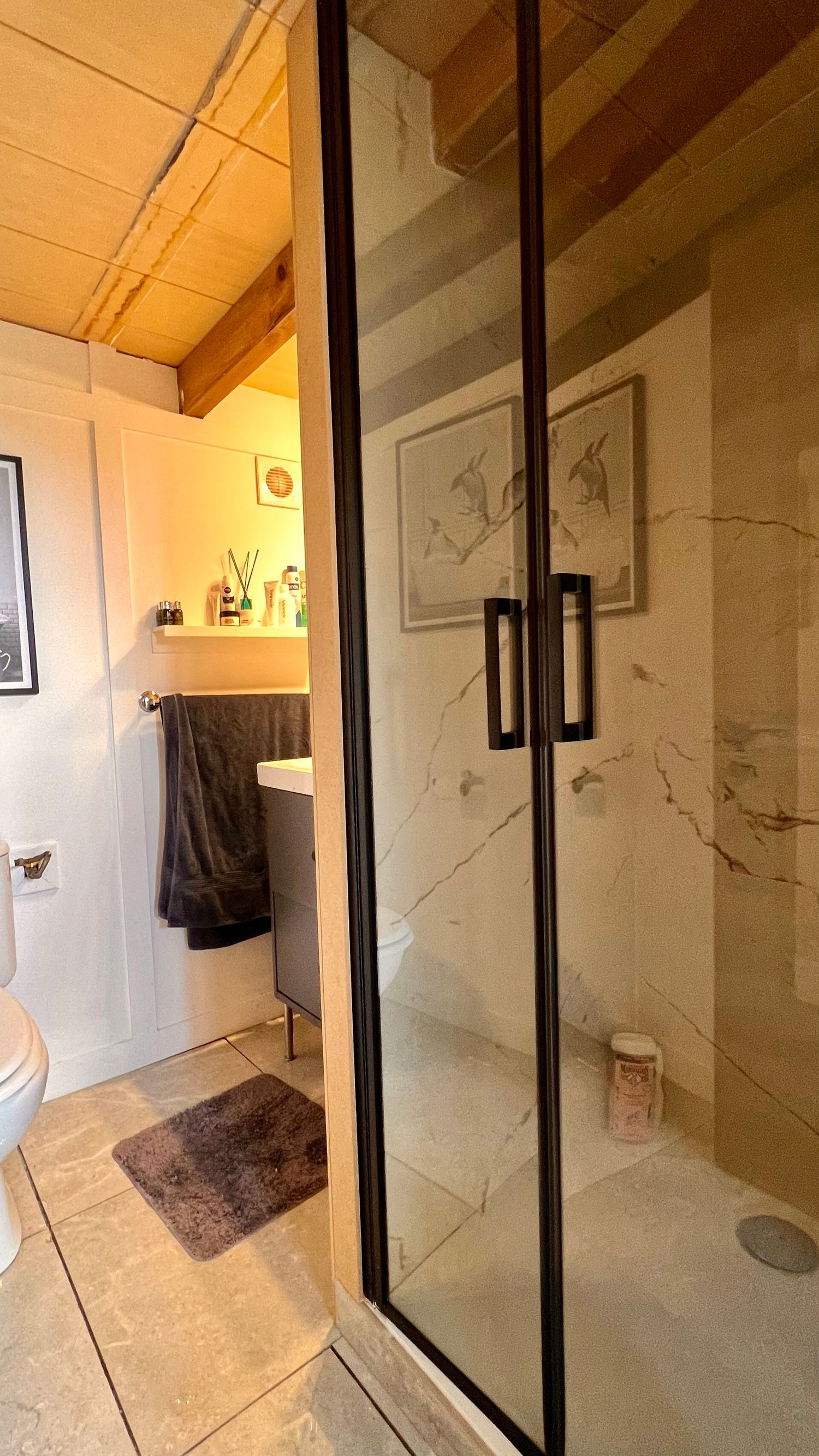 Modern bathroom interior with black-framed glass shower doors, marble-look walls, and beige floor tiles.