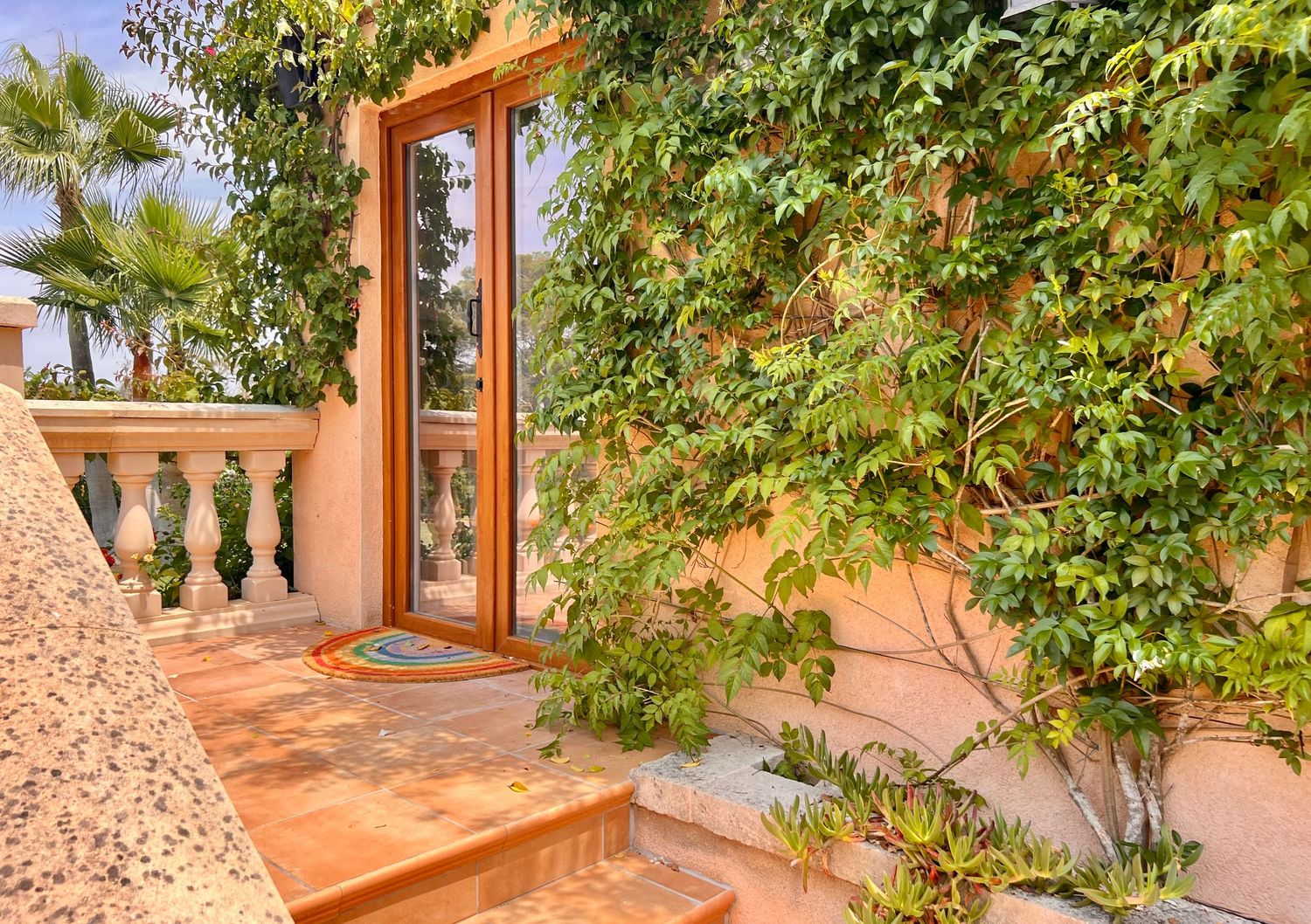 Terracotta colored Mediterranean villa entrance with wooden door, stone balustrade and lush green climbing vines.