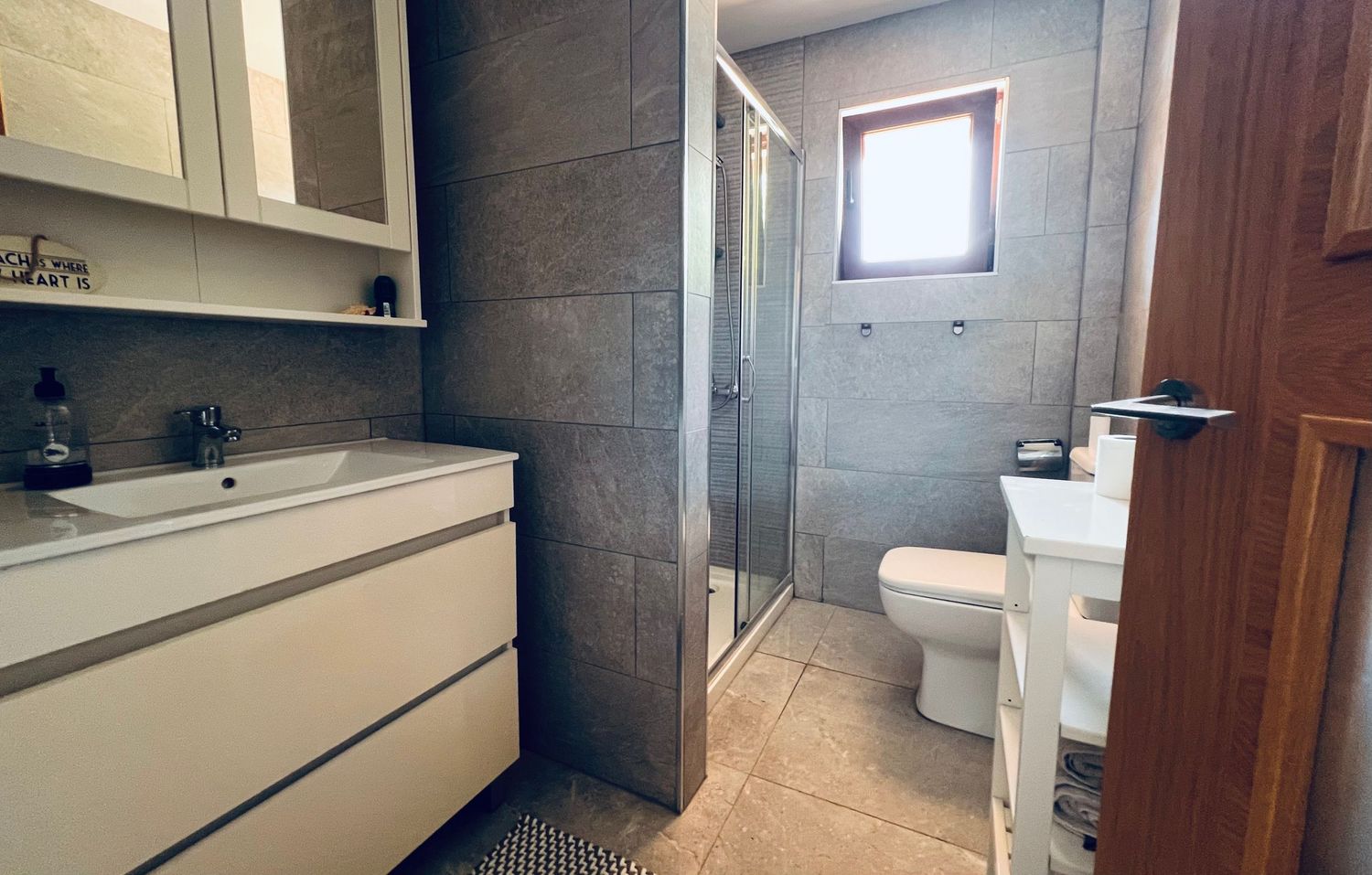 Modern bathroom interior with floating vanity, gray tile walls, glass shower enclosure and small window providing natural light.