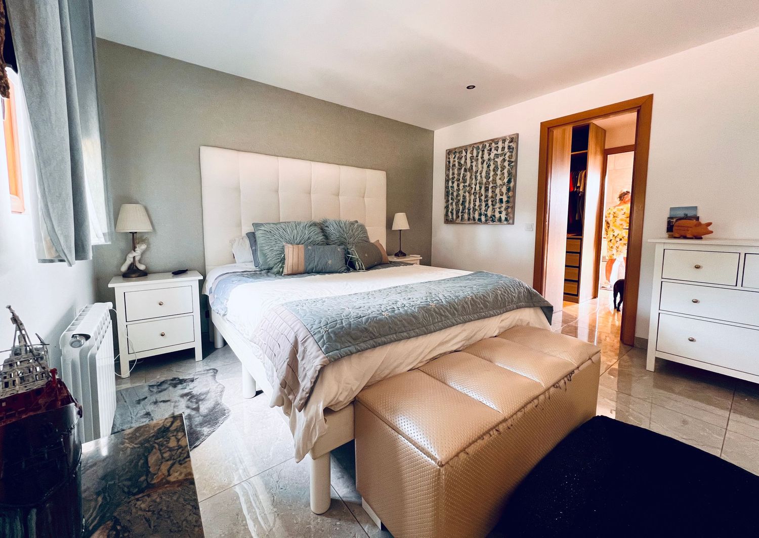Modern bedroom interior with white upholstered headboard, blue bedding, tan storage bench and marble-style flooring.