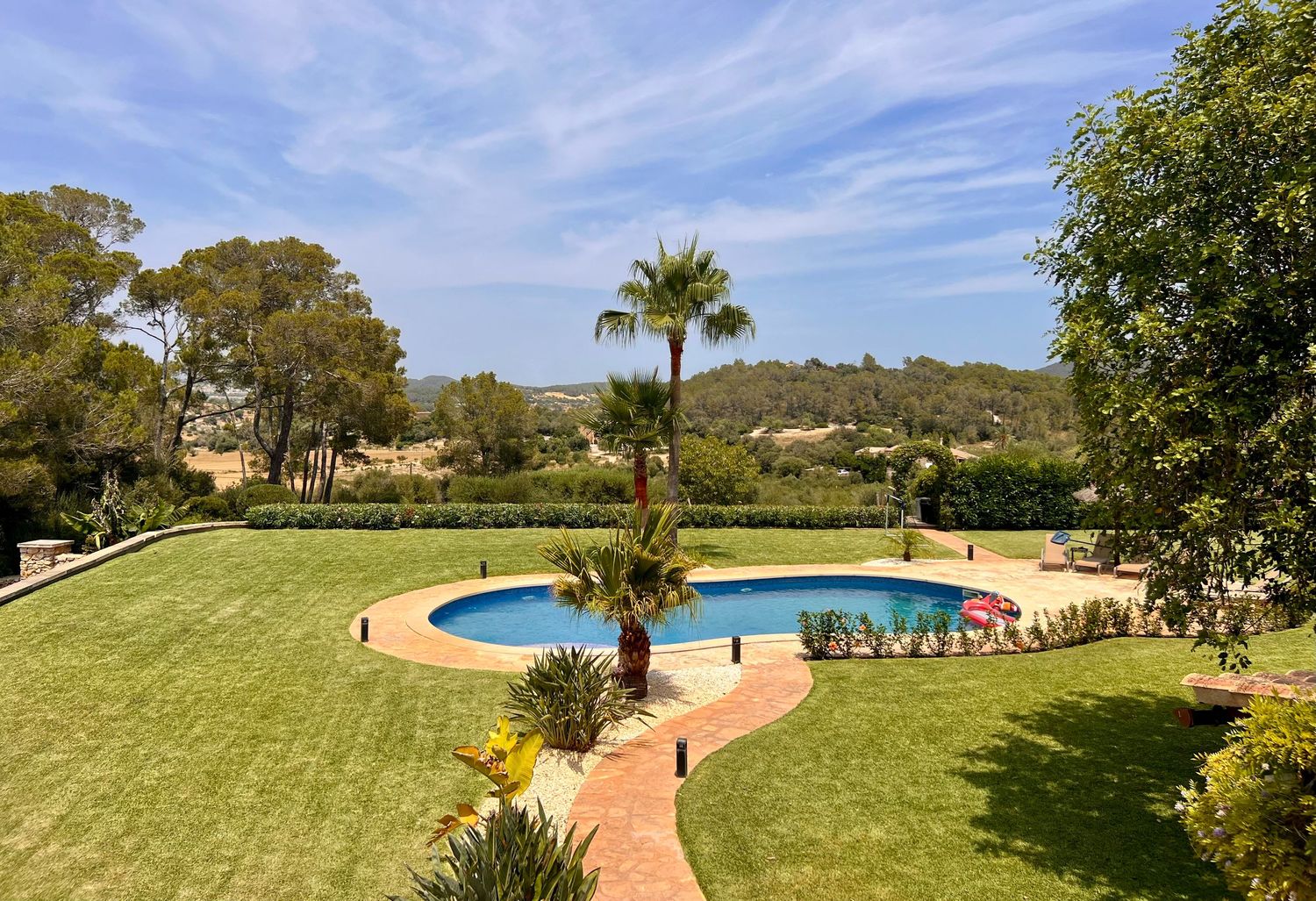 A curved swimming pool surrounded by lush green lawn and palm trees with a Spanish villa backdrop against blue sky.