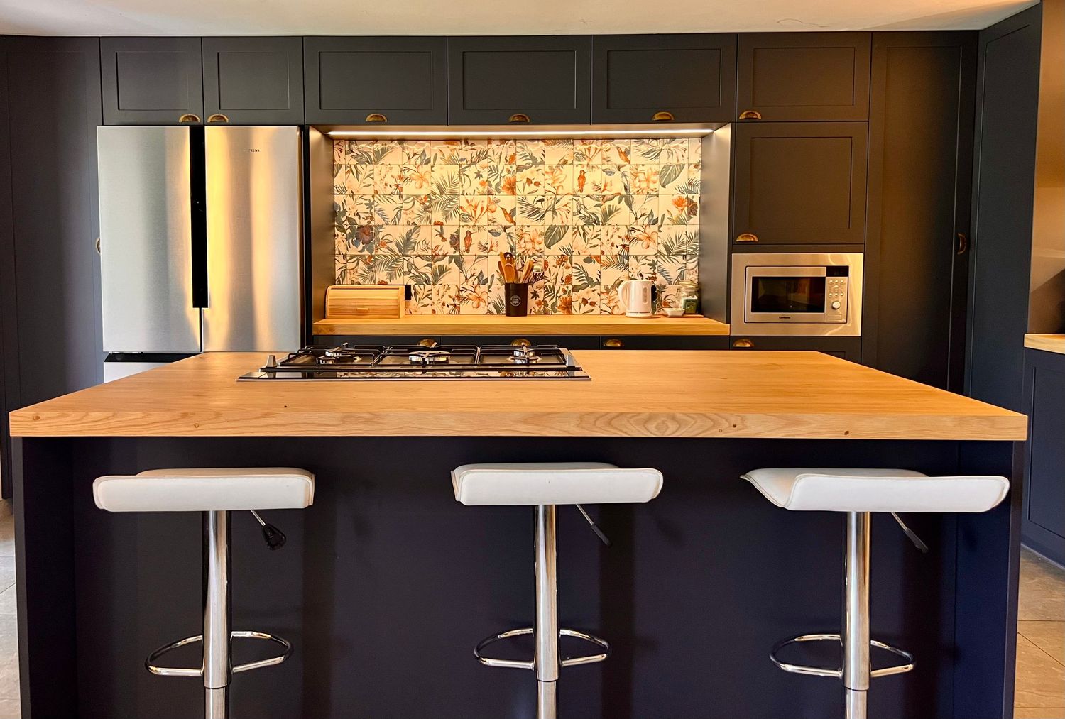 Modern kitchen with dark cabinets features a wooden breakfast bar, white barstools, and decorative floral wallpaper backsplash.