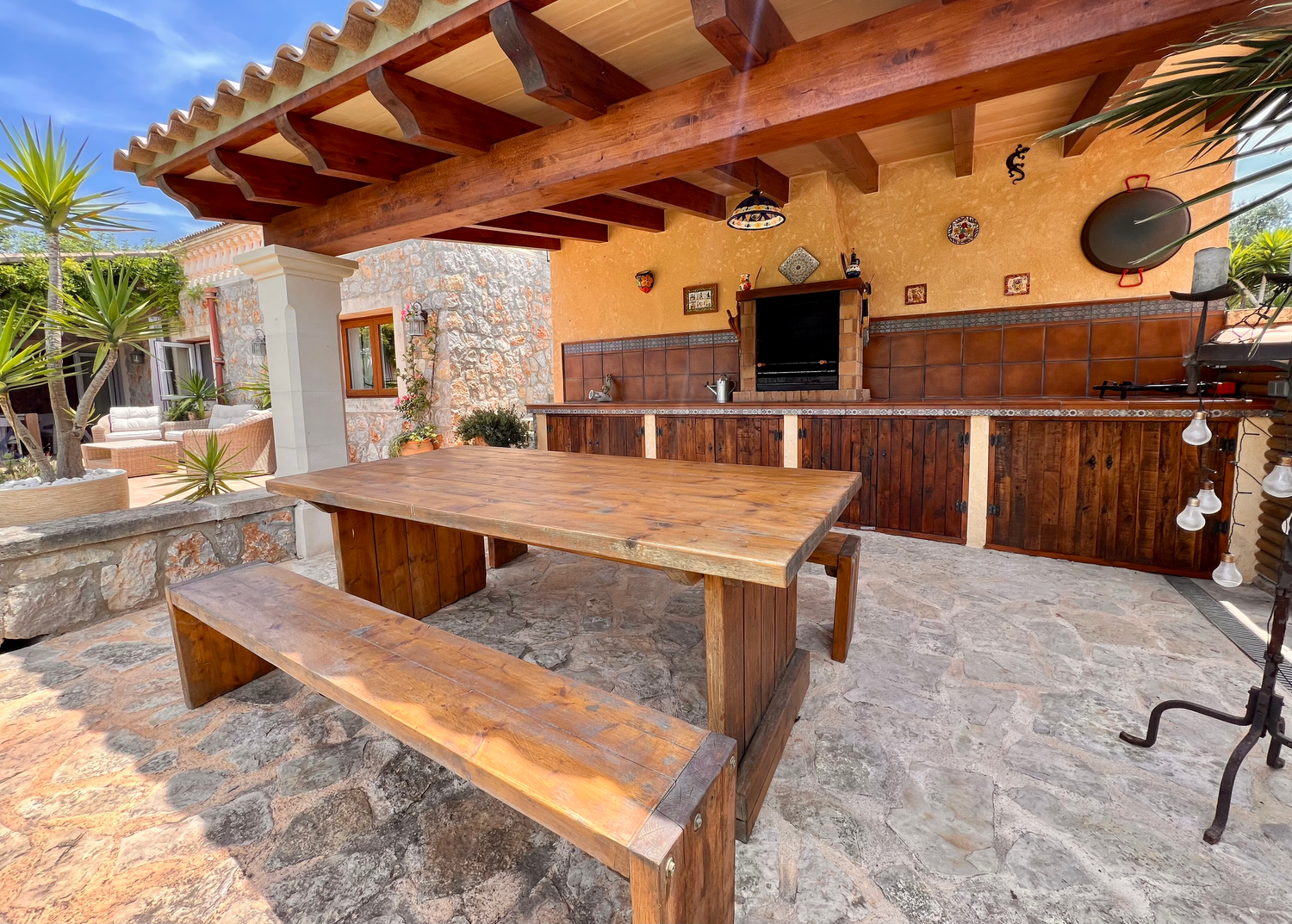 Rustic outdoor kitchen and dining area with wooden pergola, stone accents, and built-in bench seating on paved patio.