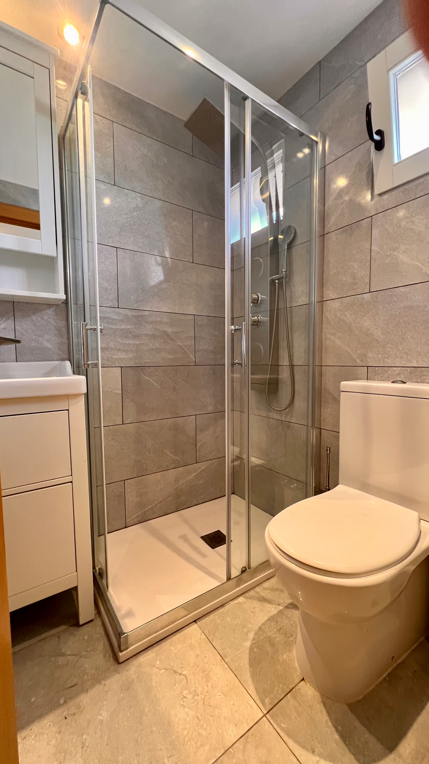 Modern bathroom featuring a glass shower enclosure, white toilet, and gray tile walls with warm lighting fixtures.