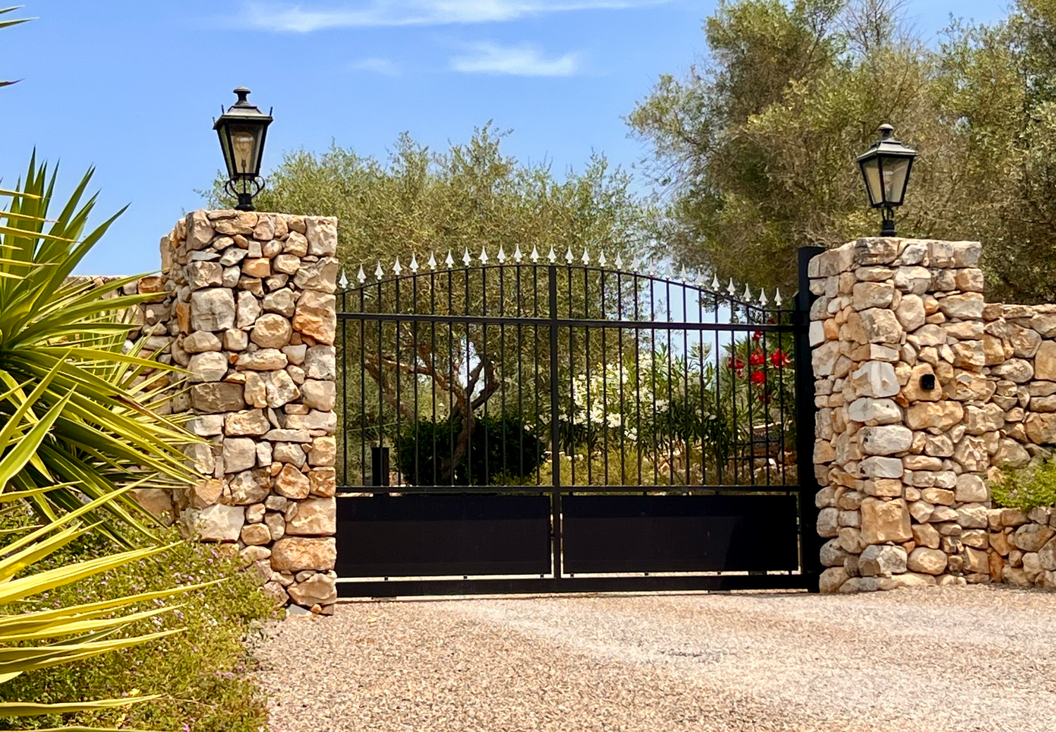 Ornate black metal driveway gate set between stone pillars with lanterns and desert landscaping.