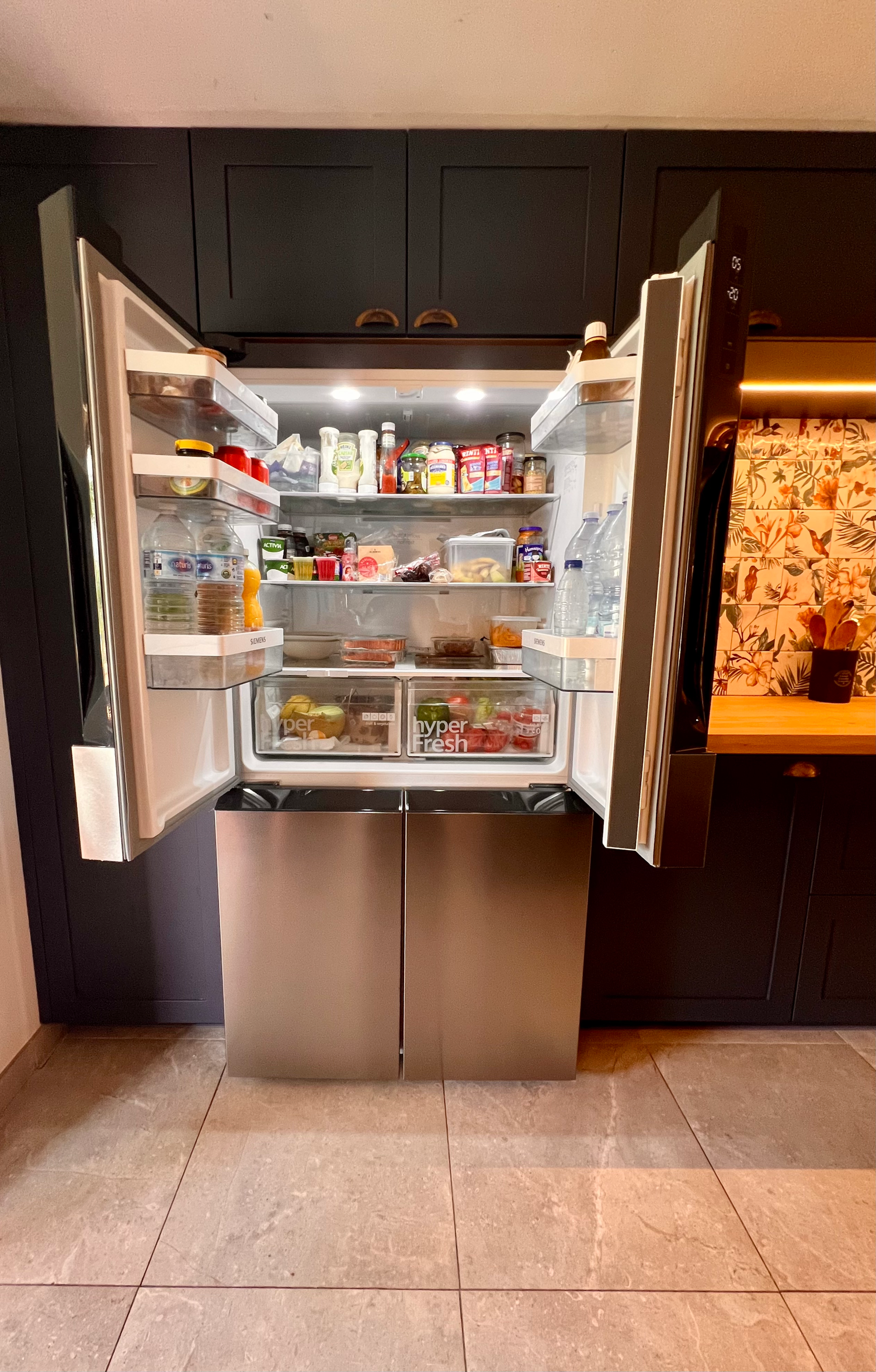 Modern stainless steel French door refrigerator with fully stocked shelves built into dark cabinetry on tiled floor.