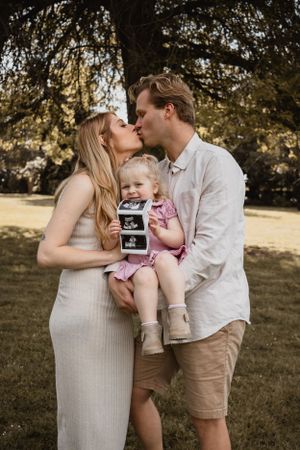 Mum and dad kissing while holding their daughter and ultrasound picture.