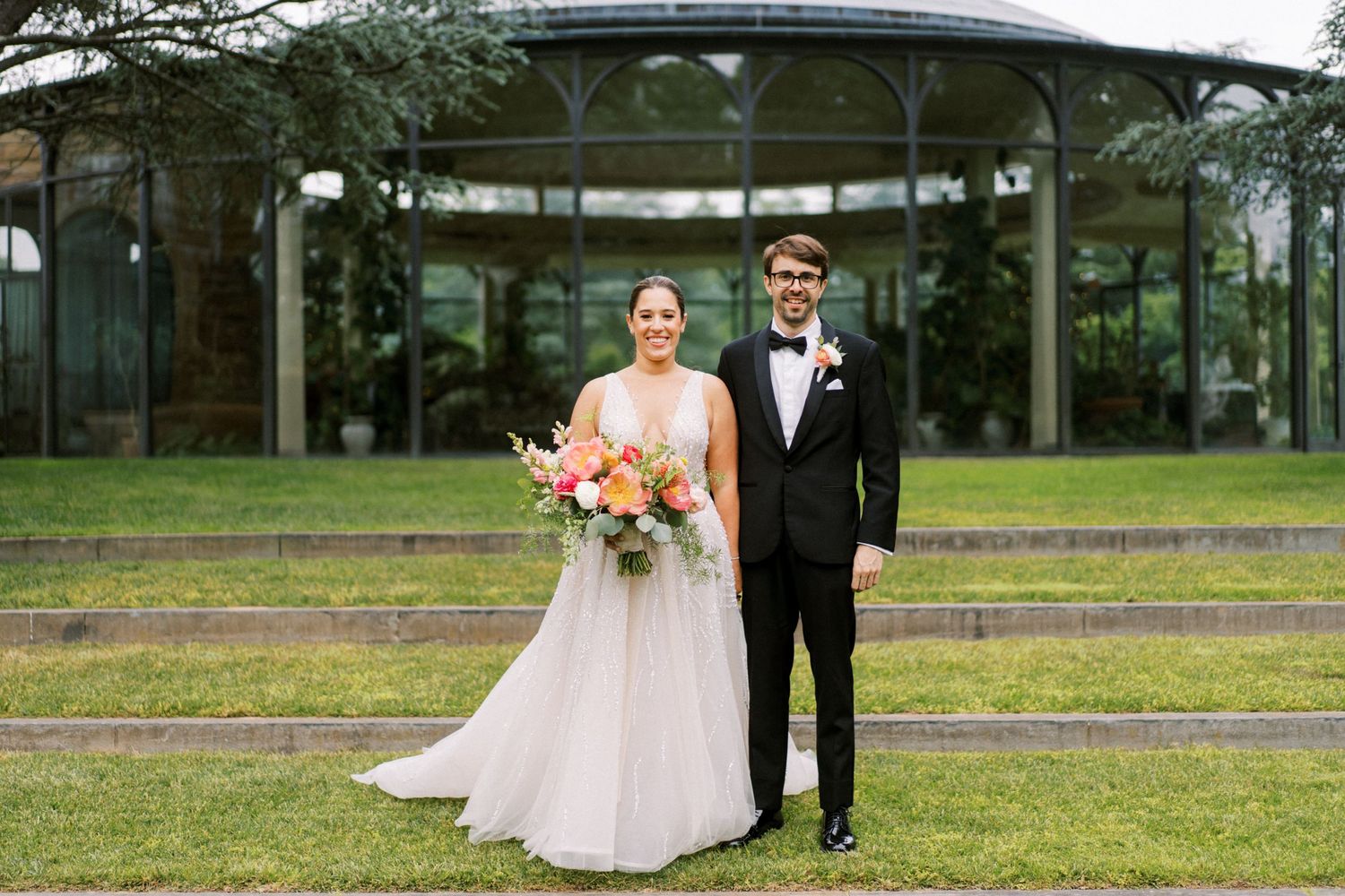 A bride and groom pose for a portrait in front of a glass conservatory building on their wedding day.