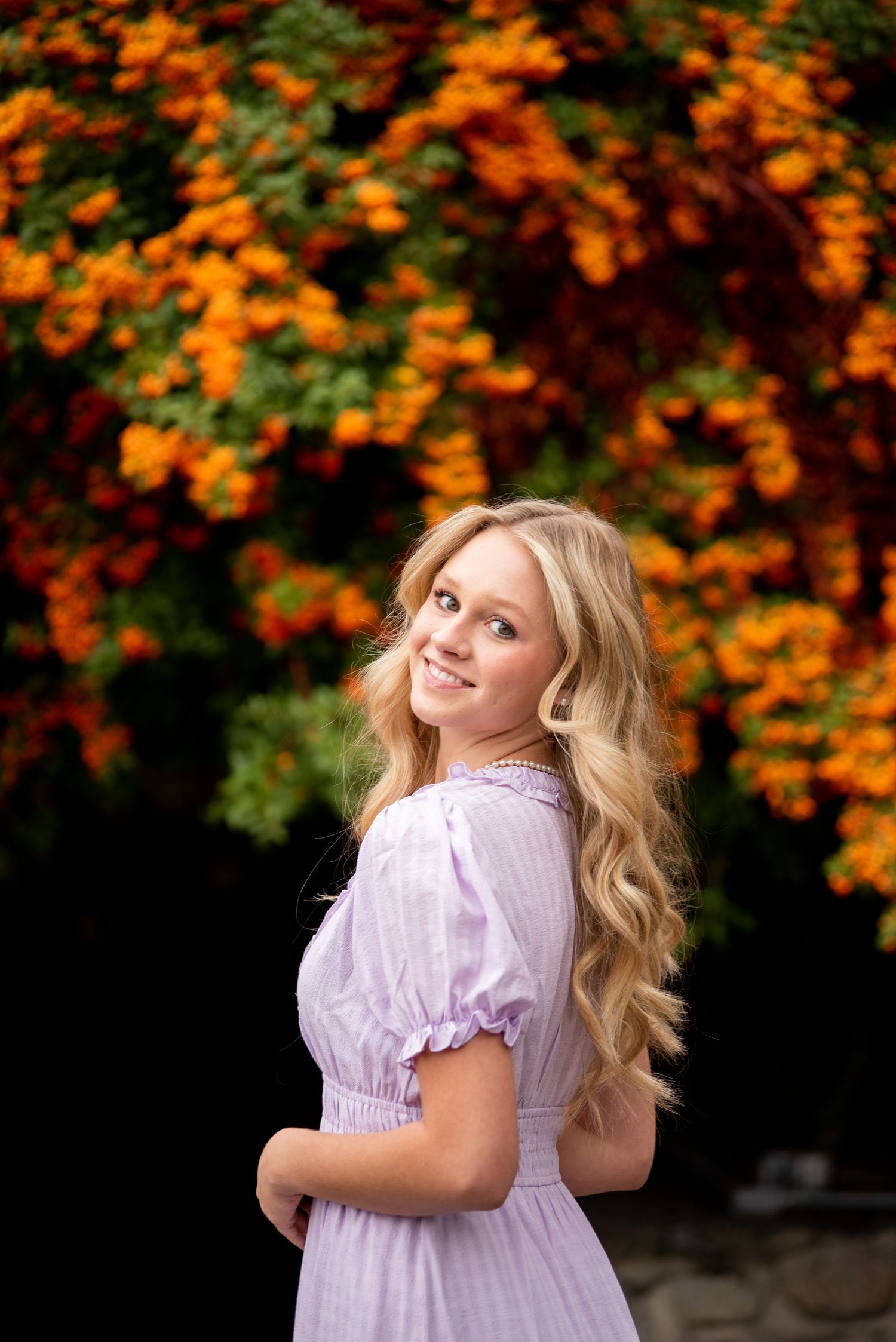 A woman in a lavender dress poses against vibrant orange fall foliage.