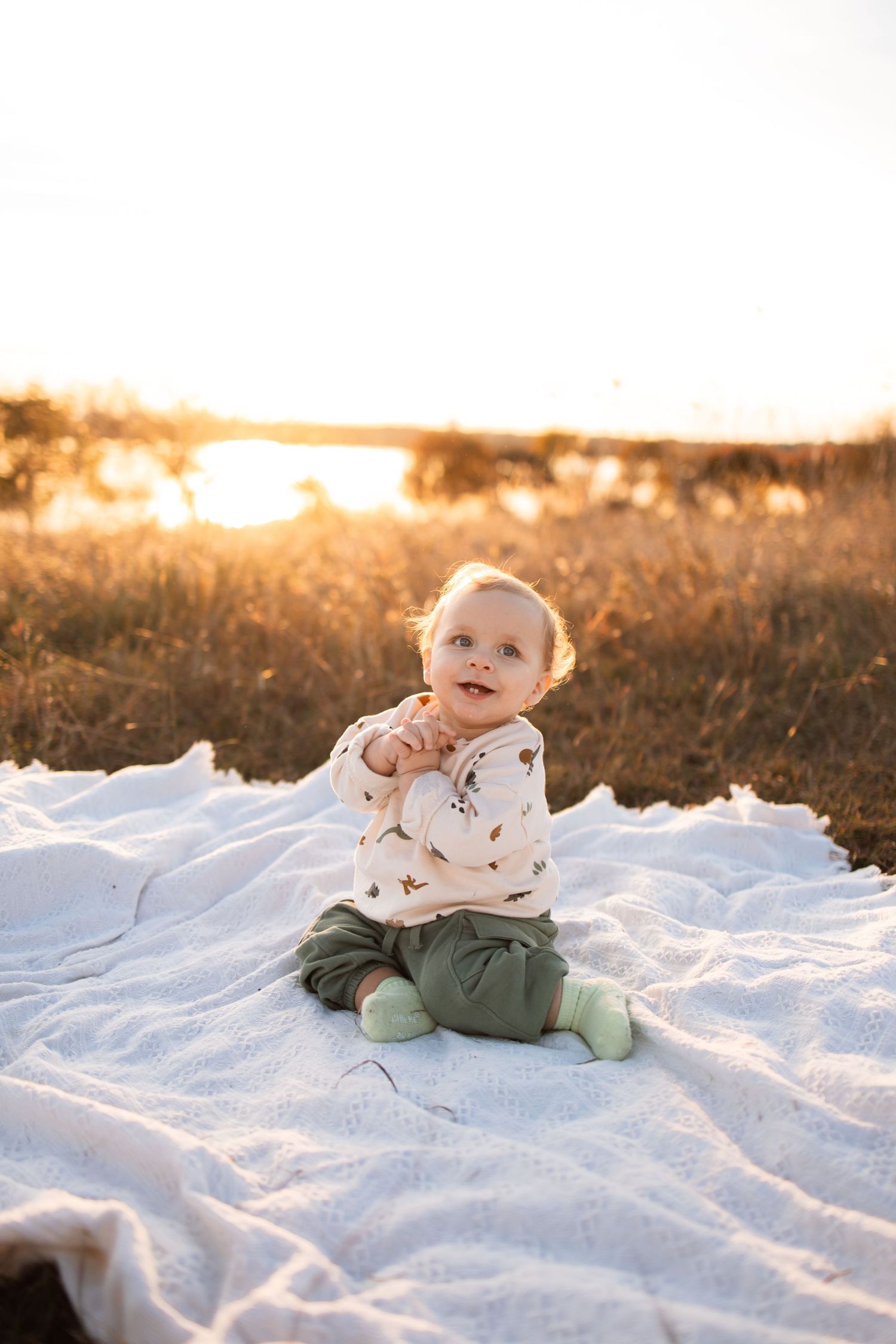 A baby sits on a white blanket outdoors during golden hour wearing a patterned sweater.