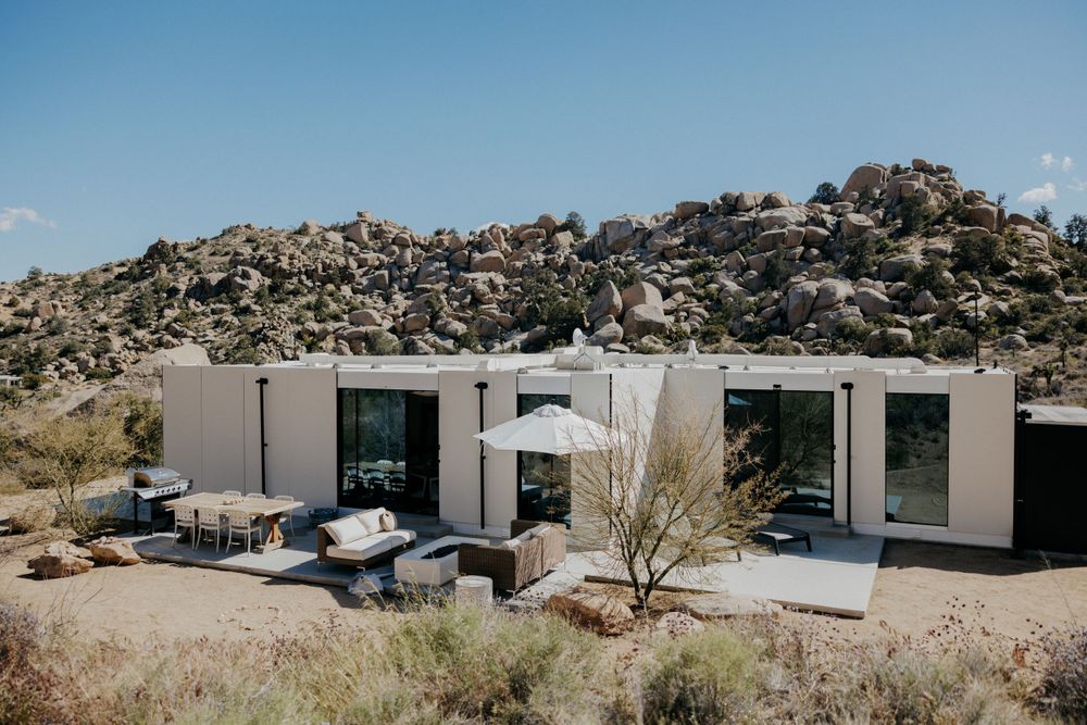 Modern minimalist house nestled against rocky desert landscape with mountain backdrop.
