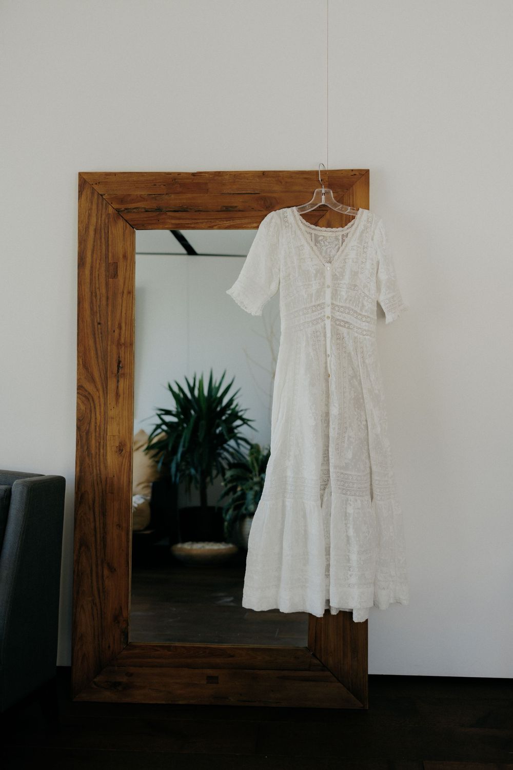 White lace vintage dress hanging on wooden framed mirror with indoor plant reflection.