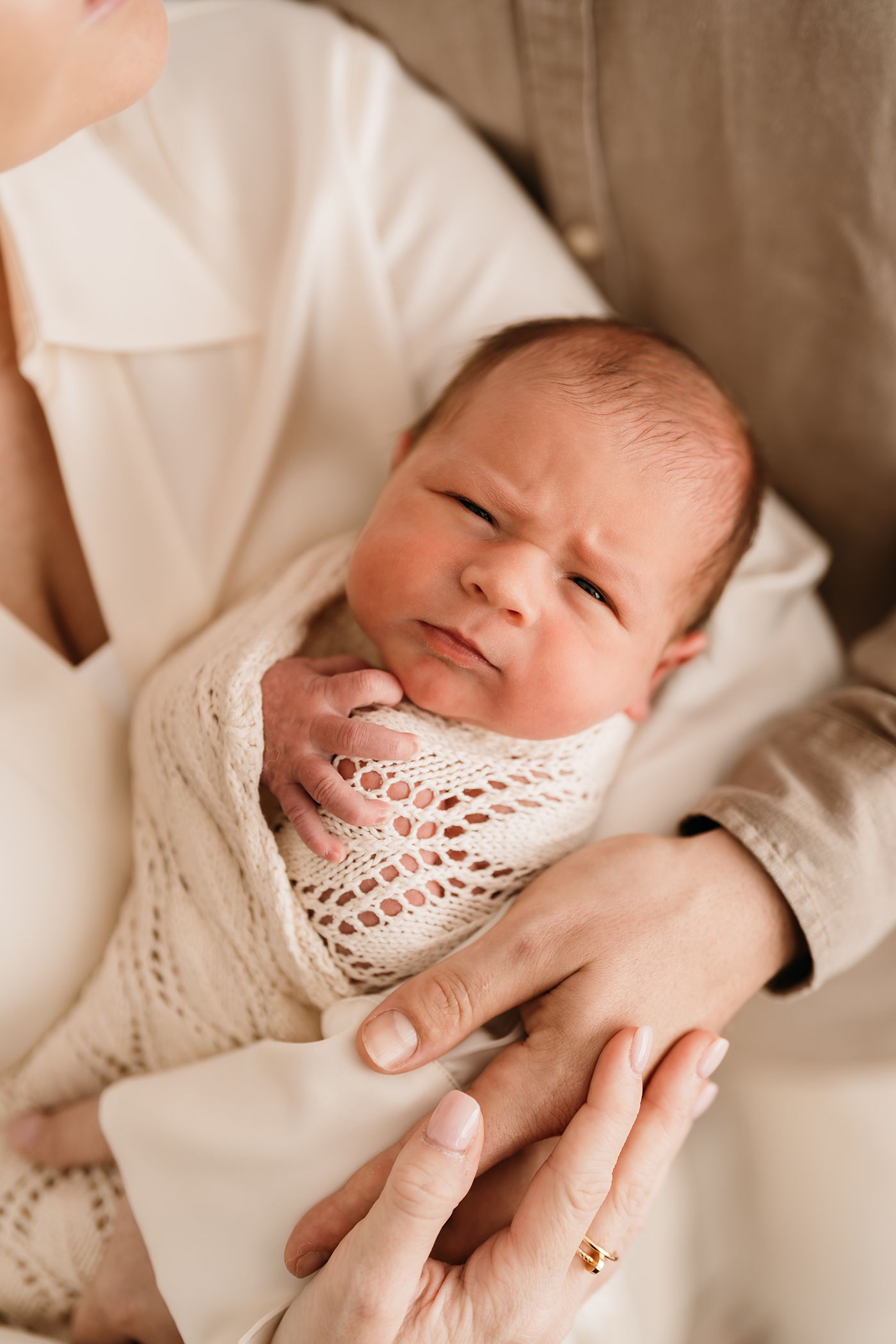 Natural newborn photos. A newborn baby wrapped in a cream knit blanket makes an expressive face while being held in gentle hands.