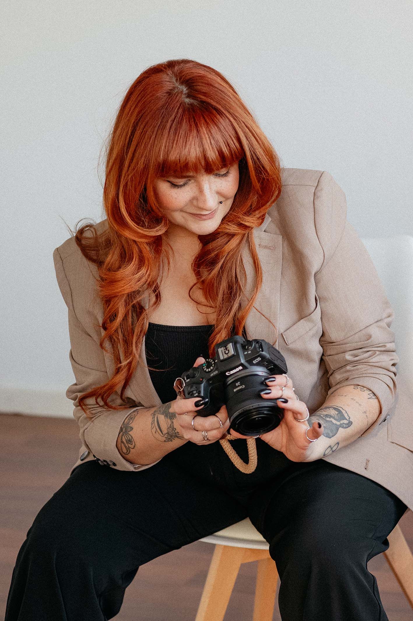 A stylish photographer wearing a beige blazer checks settings on a vintage camera while sitting on a wooden stool.