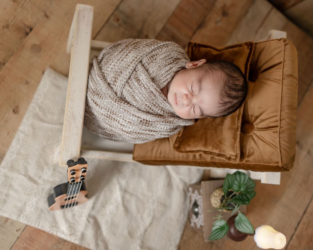 Sleeping newborn wrapped in knit blanket on brown cushion with toys and decor.