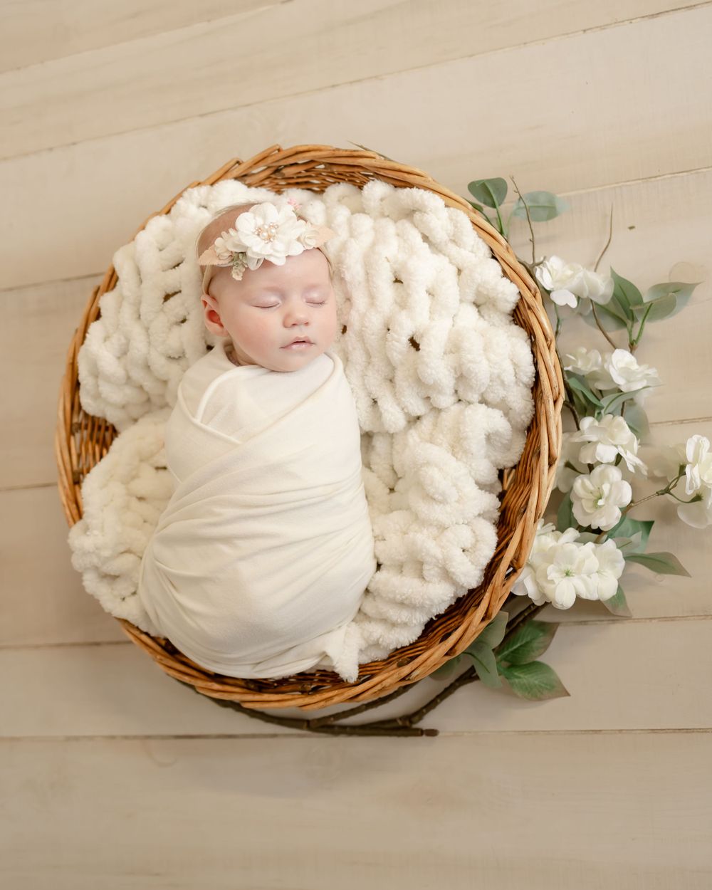 A sleeping newborn wrapped in white lies in a wicker basket with delicate floral decorations.