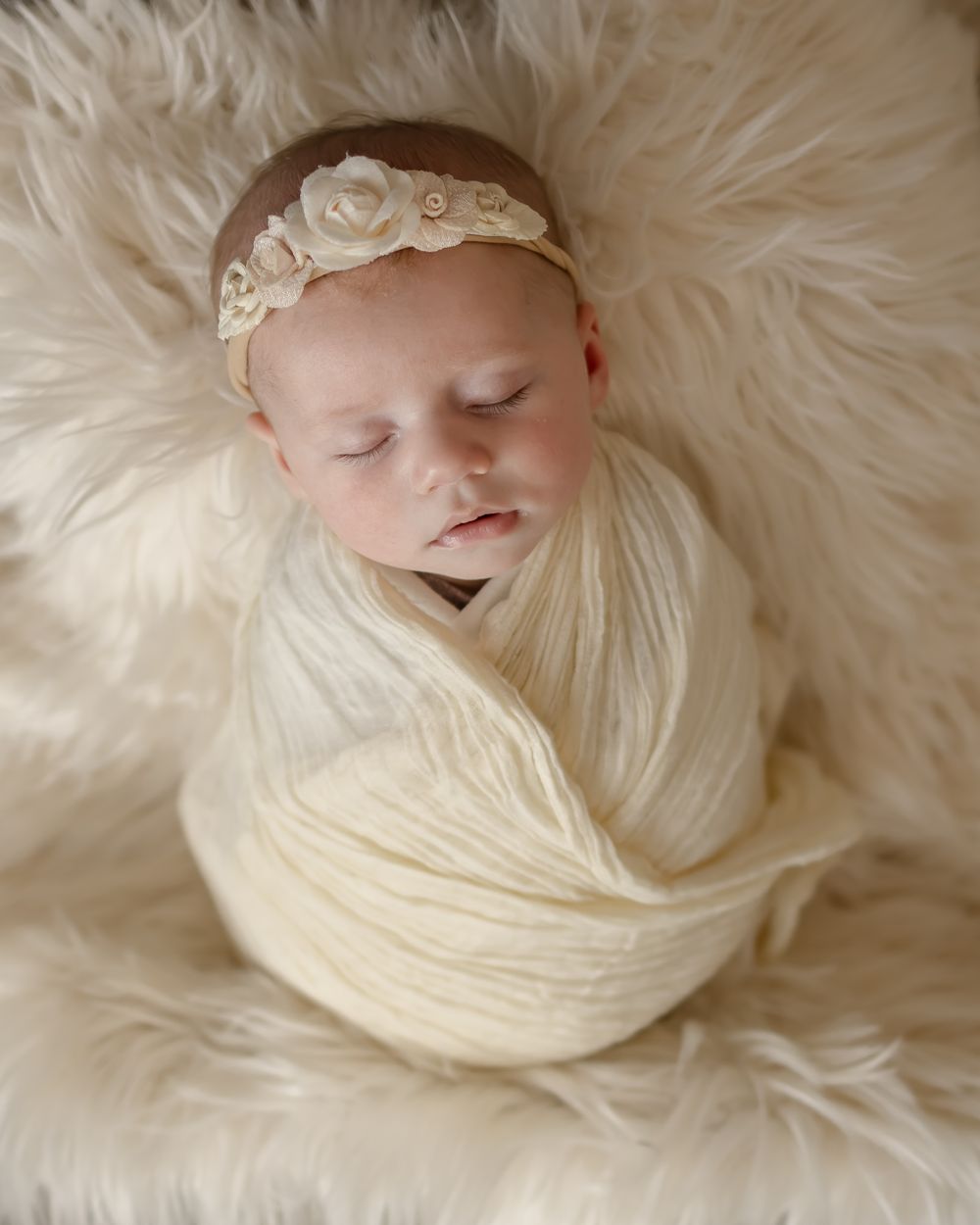 A newborn wrapped in cream fabric wearing a floral headband rests on soft white fur.