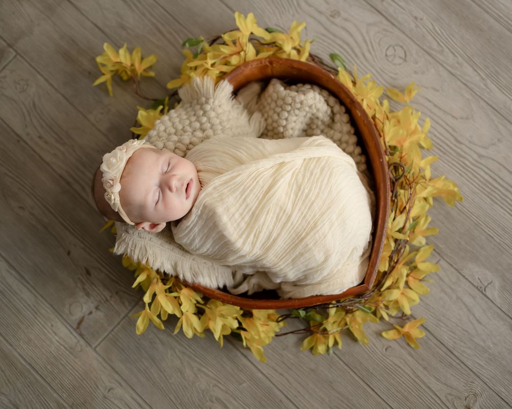 A peaceful sleeping baby wrapped in white lies in a basket surrounded by yellow daffodils.