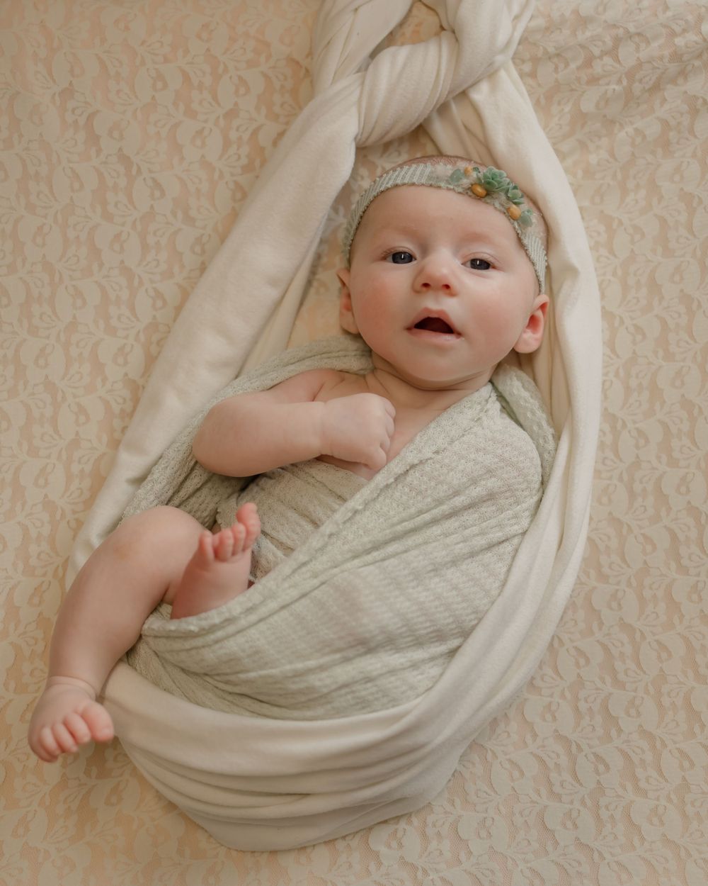 A baby wrapped in light fabric with a headband lies awake on a textured beige background.