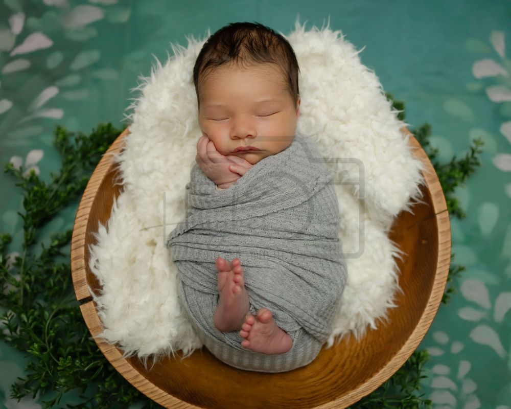 Newborn sleeping peacefully in wooden bowl on fluffy white sheepskin with green foliage backdrop.
