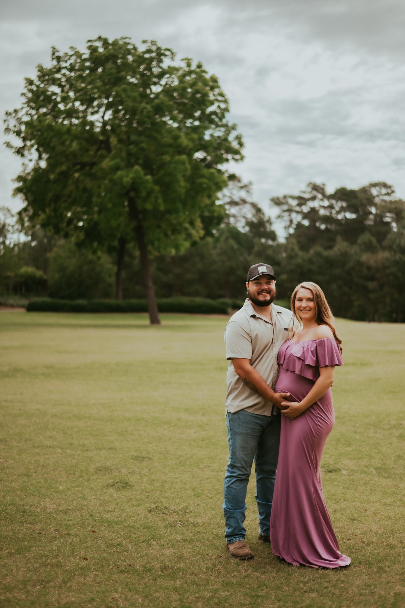 Outdoor maternity portrait of couple in casual attire standing in grassy field.