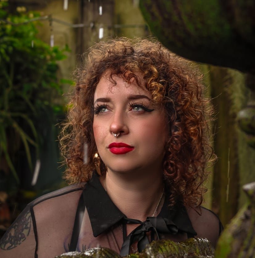 Artistic outdoor portrait of a woman with vibrant red curly hair and red lipstick in a dark outfit against a forest backdrop.