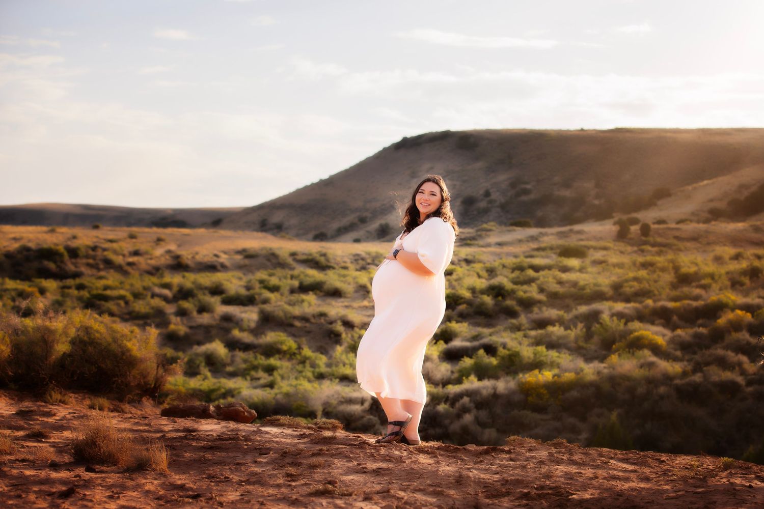 Woman in flowing white dress poses on red rocks during sunset maternity photo session.