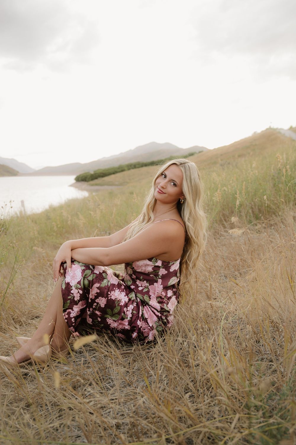 Blonde woman in floral dress sitting in golden grass near a lake with mountains in the background.