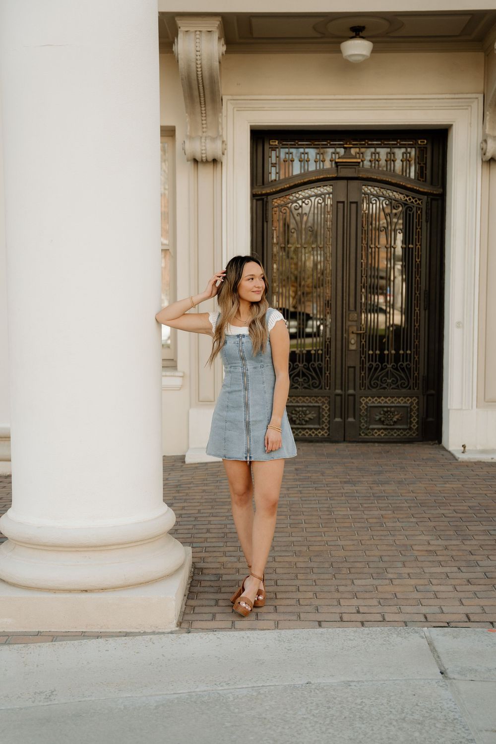 Woman in denim dress poses against elegant building entrance with ornate door and column.