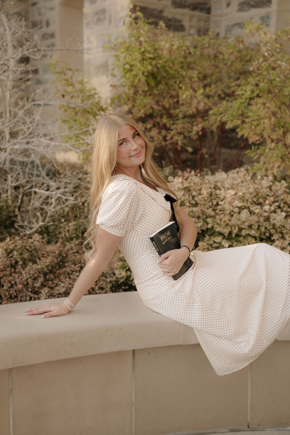 Young blonde woman in white polka dot dress sitting outdoors holding a Bible, smiling gracefully.