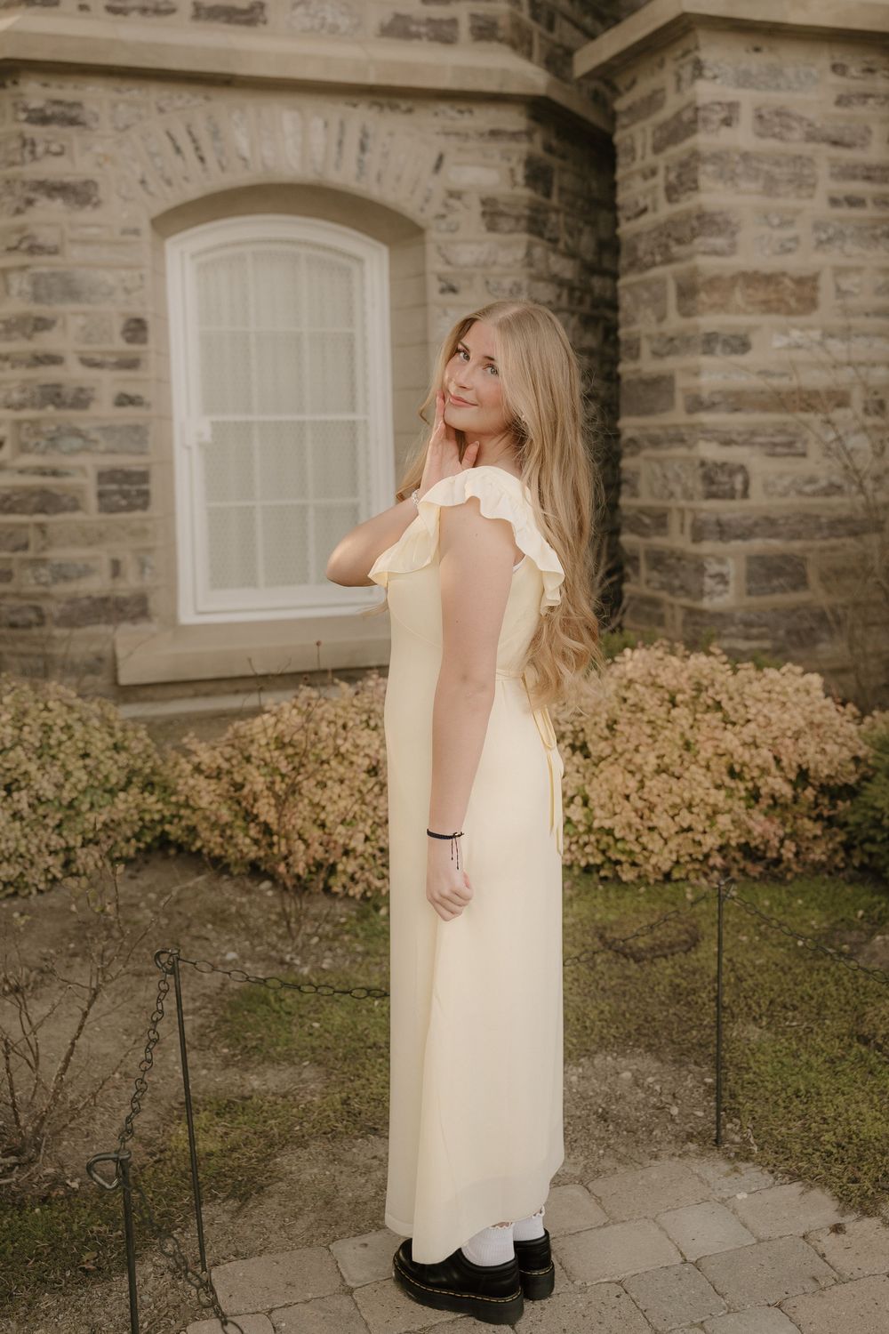 Young woman in a pale yellow maxi dress with ruffle sleeves posing outdoors near a stone building.