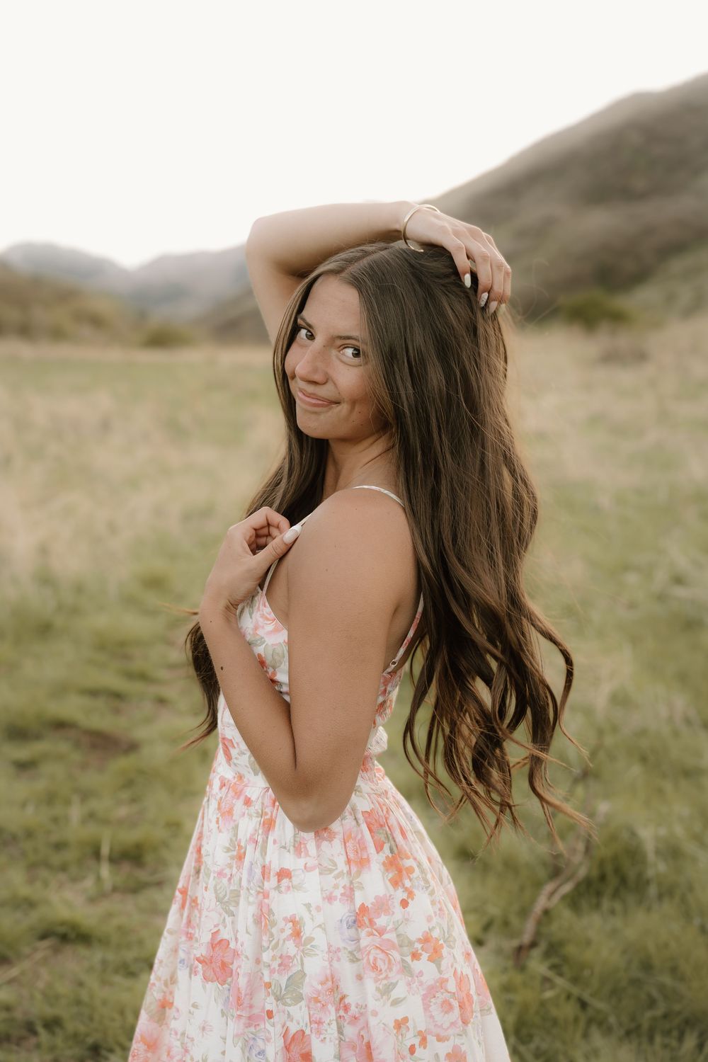 Young woman in floral sundress posing in green meadow with mountains, long brunette hair, smiling over shoulder.