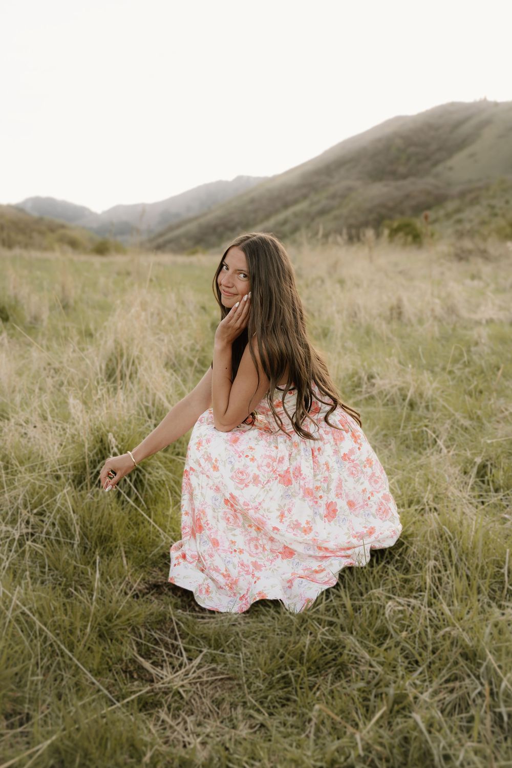 Young woman in floral maxi dress sitting in grassy meadow with mountains in background.