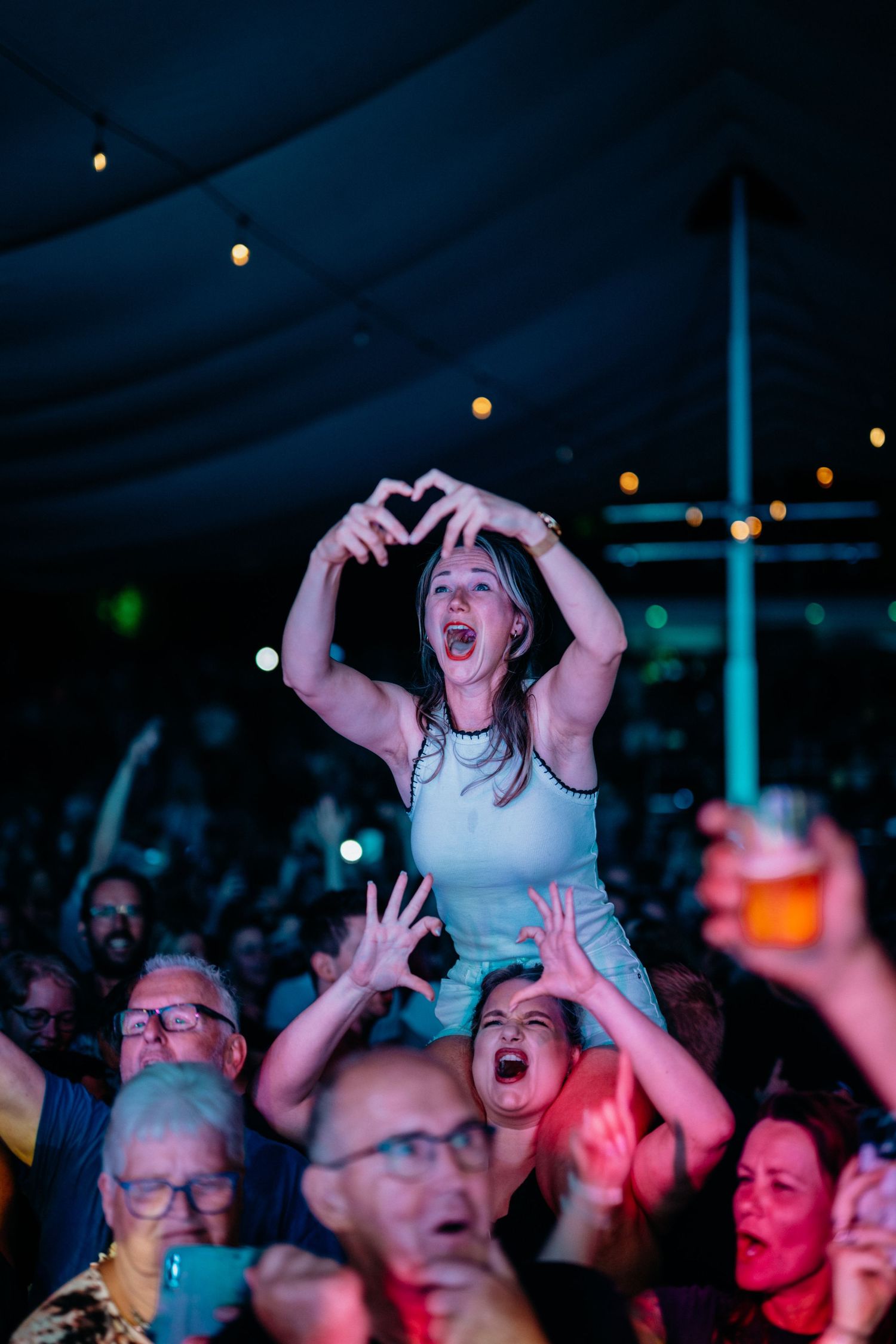 Een vrouw zit tijdens een concert op de schouders van haar vriendin. Beide maken met hun vingers een hartje naar de band.
