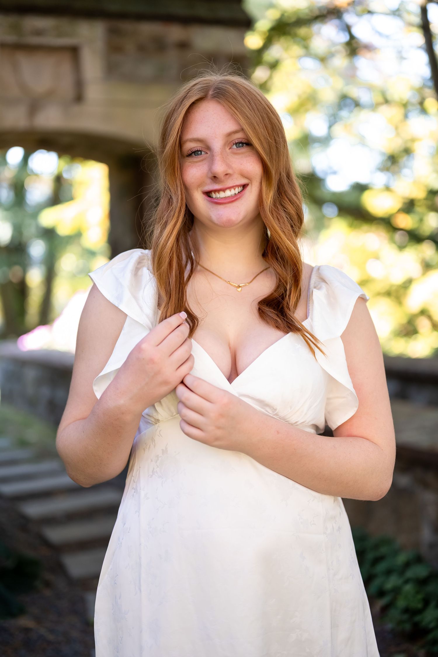 A person in a white dress poses outdoors against a blurred natural background in warm sunlight for her senior session in Masachusetts.