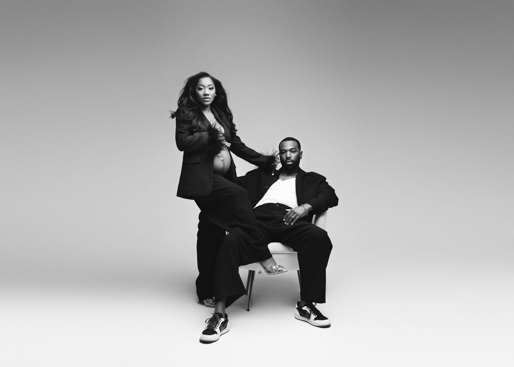 Black and white studio portrait of two people in coordinated dark outfits posing against minimal background.