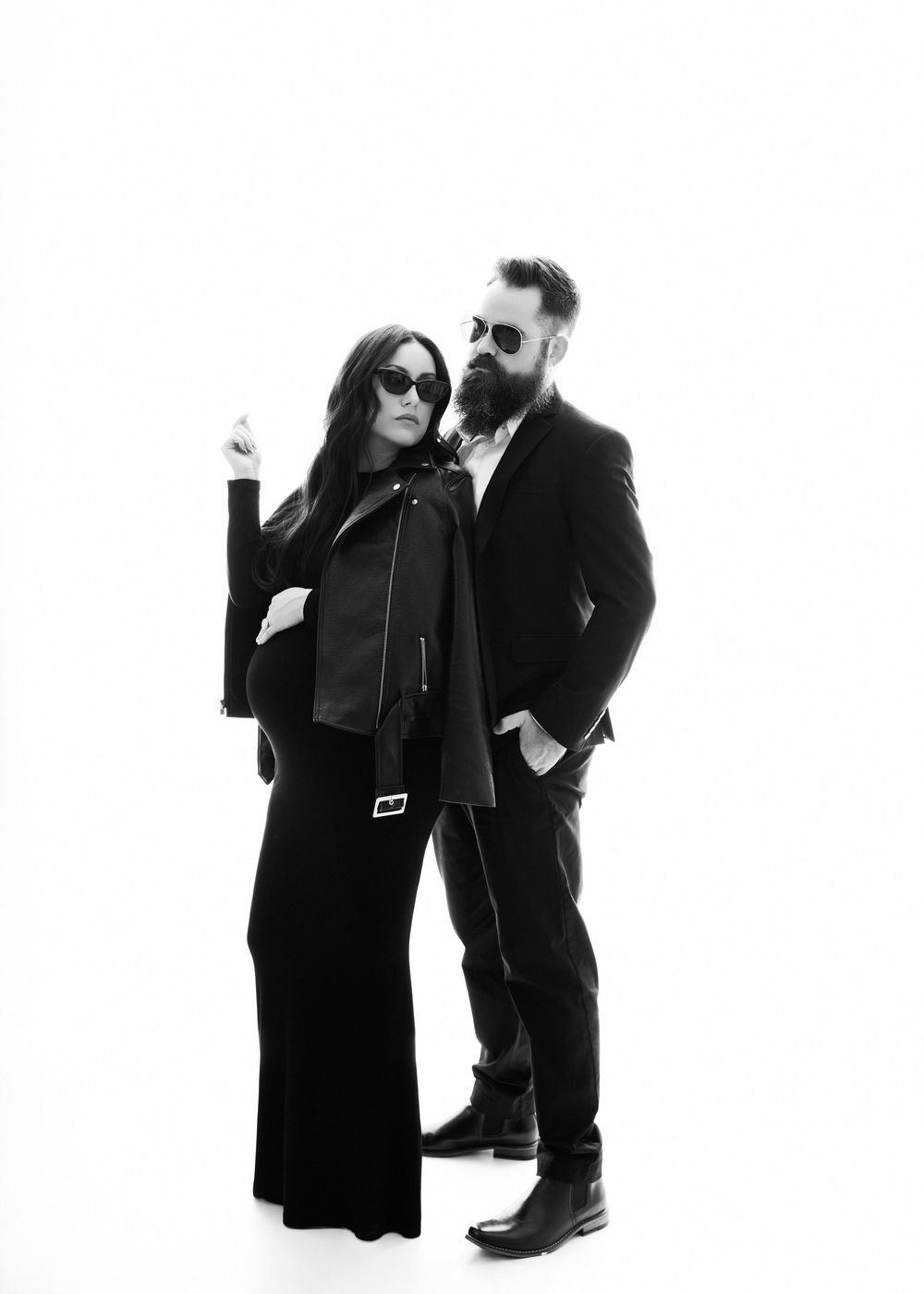 Stylish black and white portrait of couple in all-black outfits posing against white background.