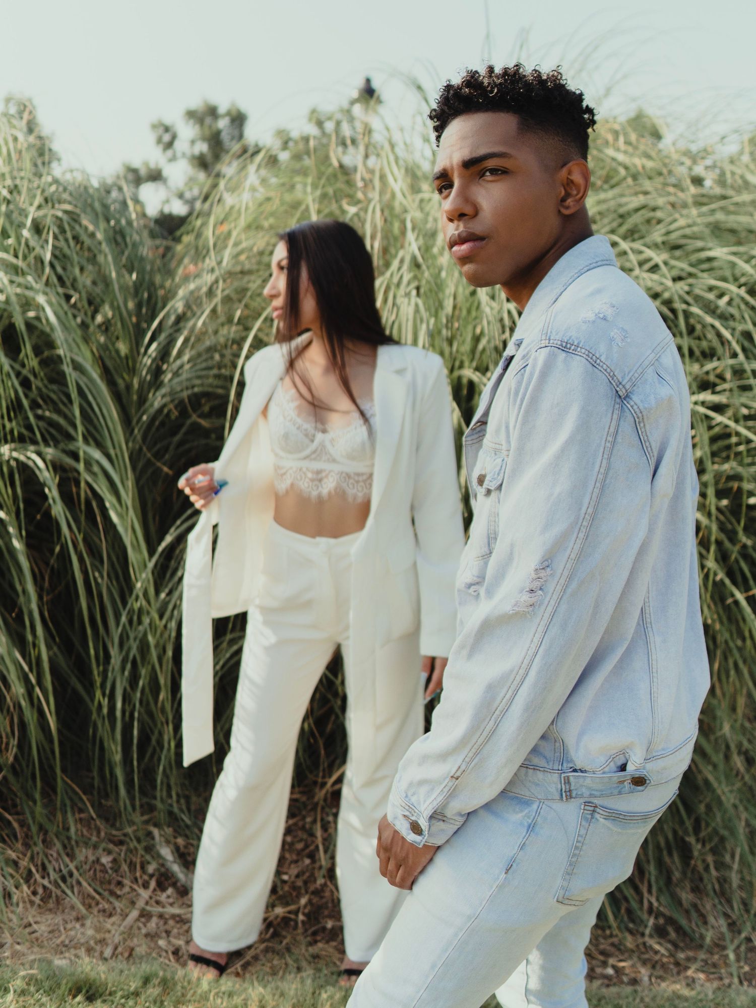 A stylish couple poses in matching white and denim outfits against tall grass.