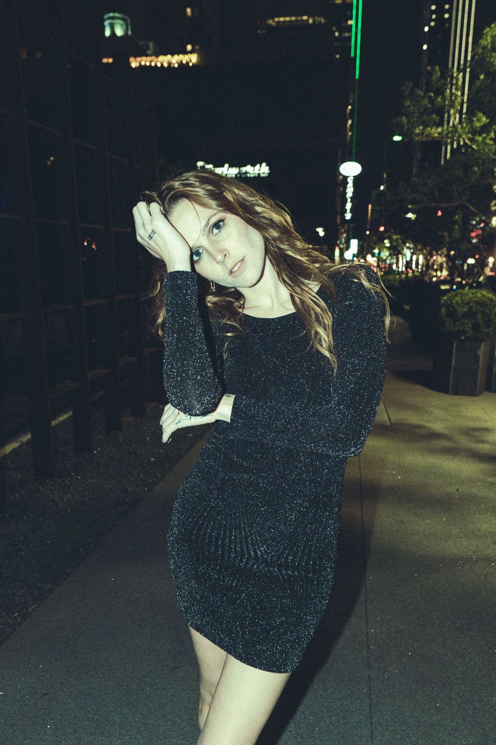 Woman in sparkly black mini dress poses on city street at night.