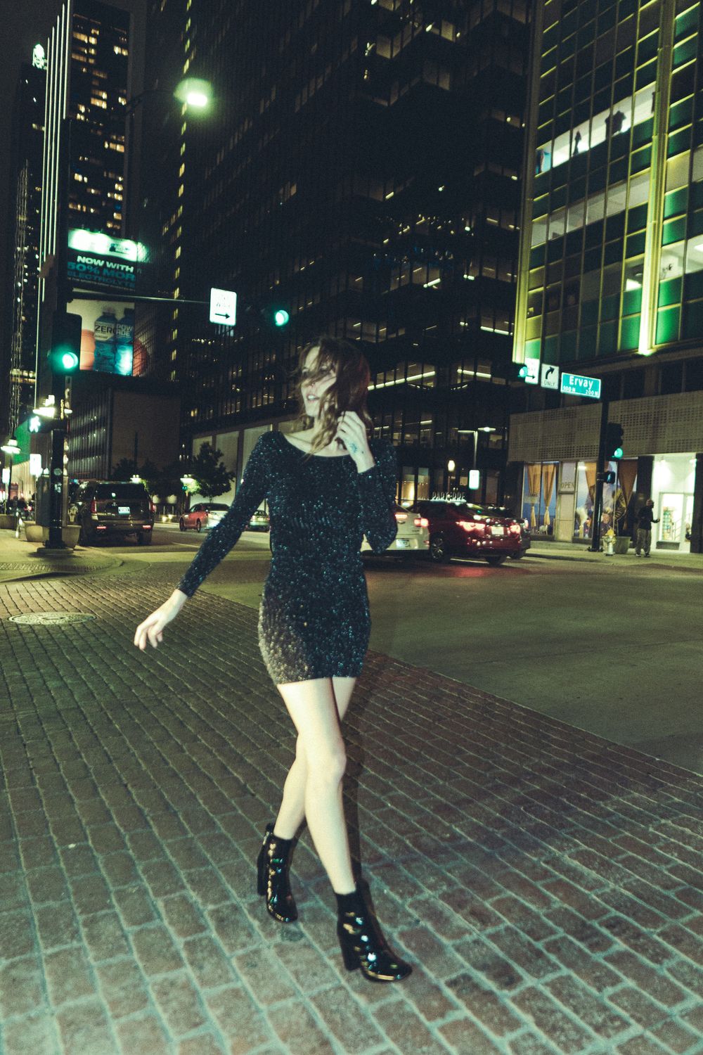 Model in short black dress poses on brick sidewalk with city lights behind.