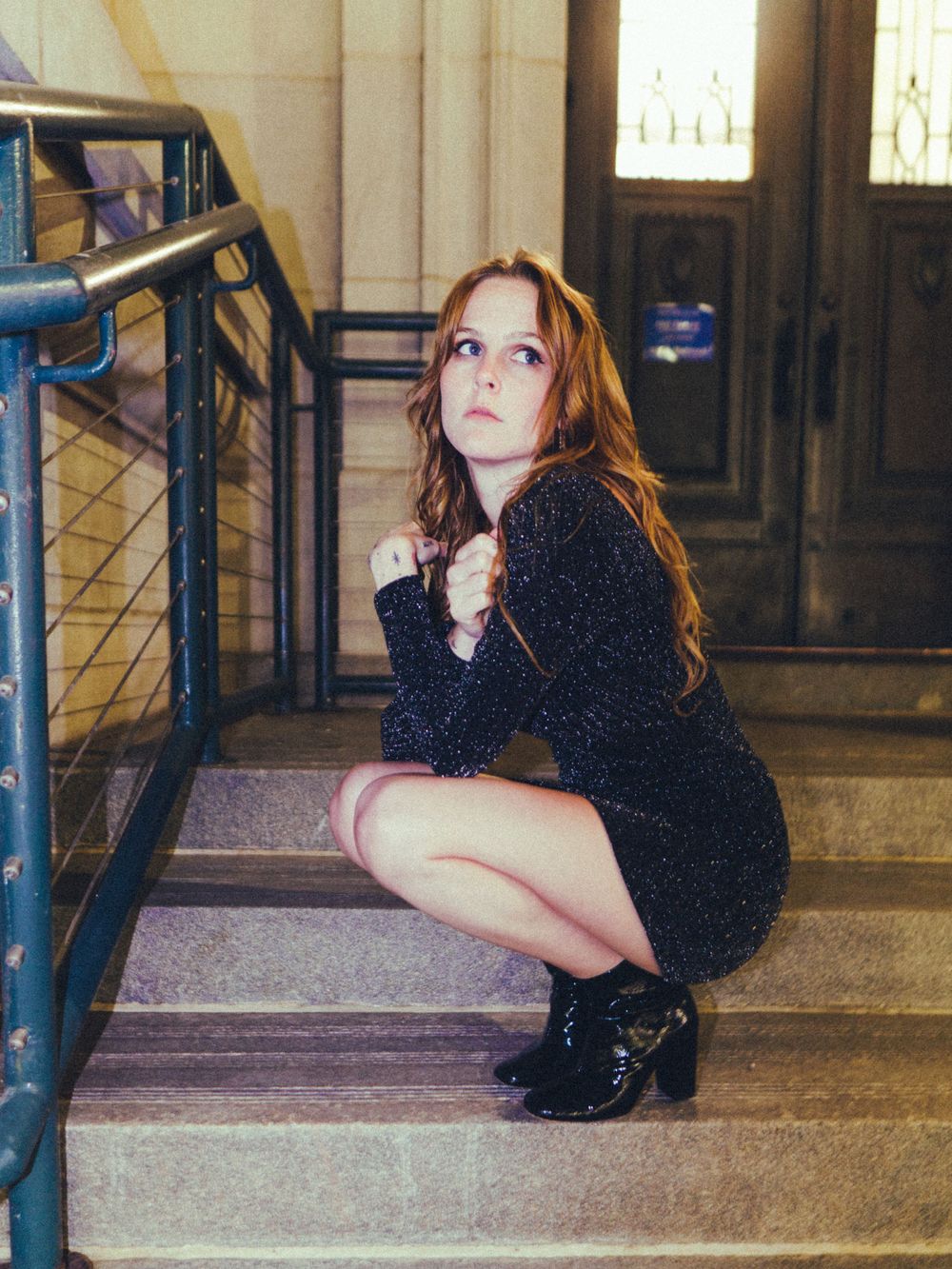 A figure in a dark sparkly dress crouches on marble stairs near a metal railing.