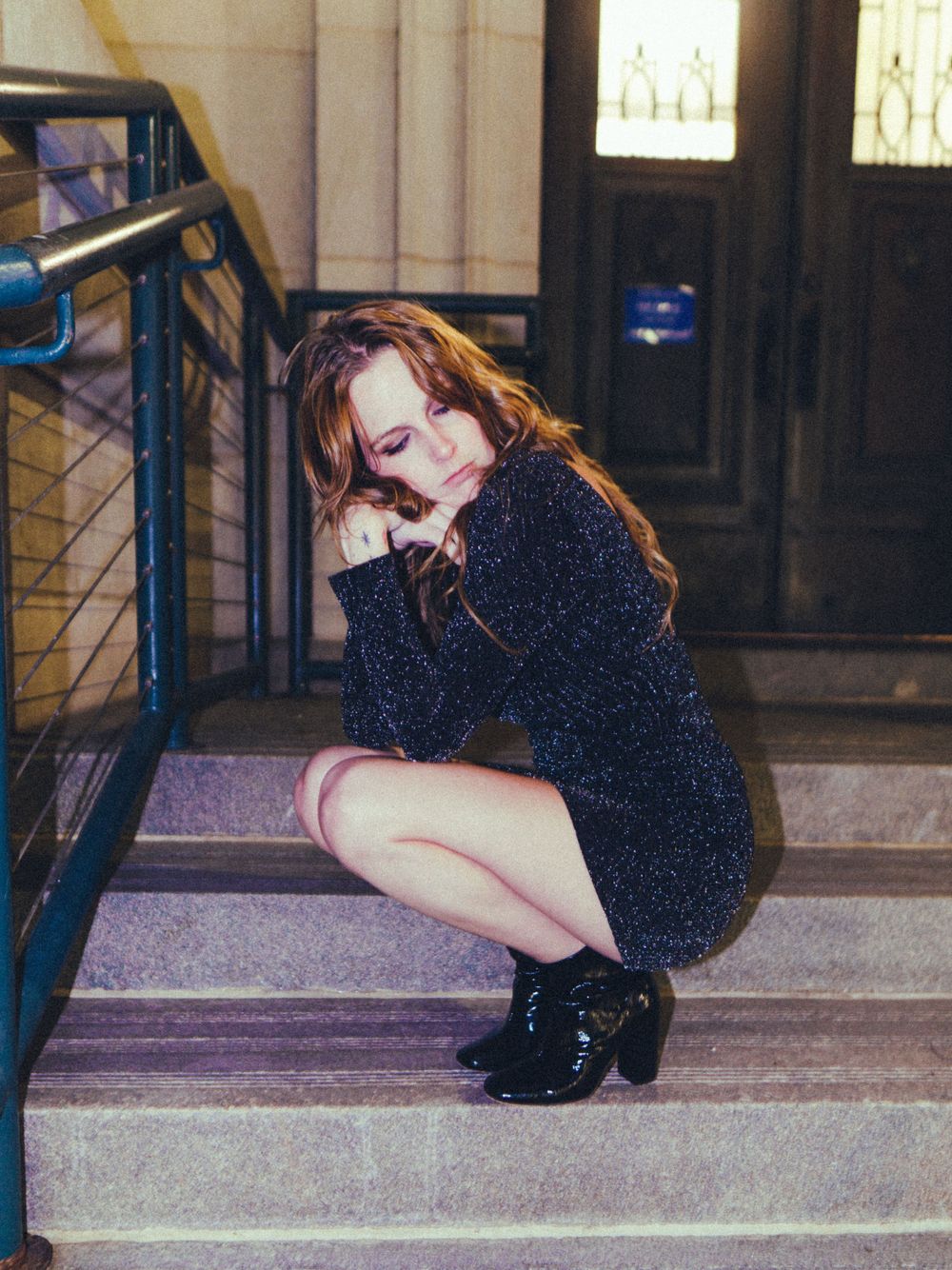 A figure in a shimmering black dress poses on stone steps indoors.
