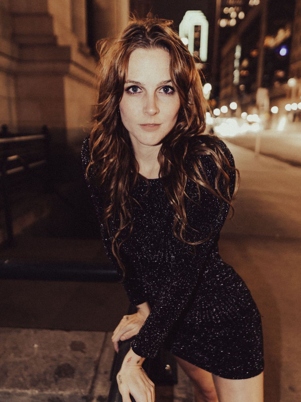 A figure in a sparkly black dress poses against a city backdrop at night.