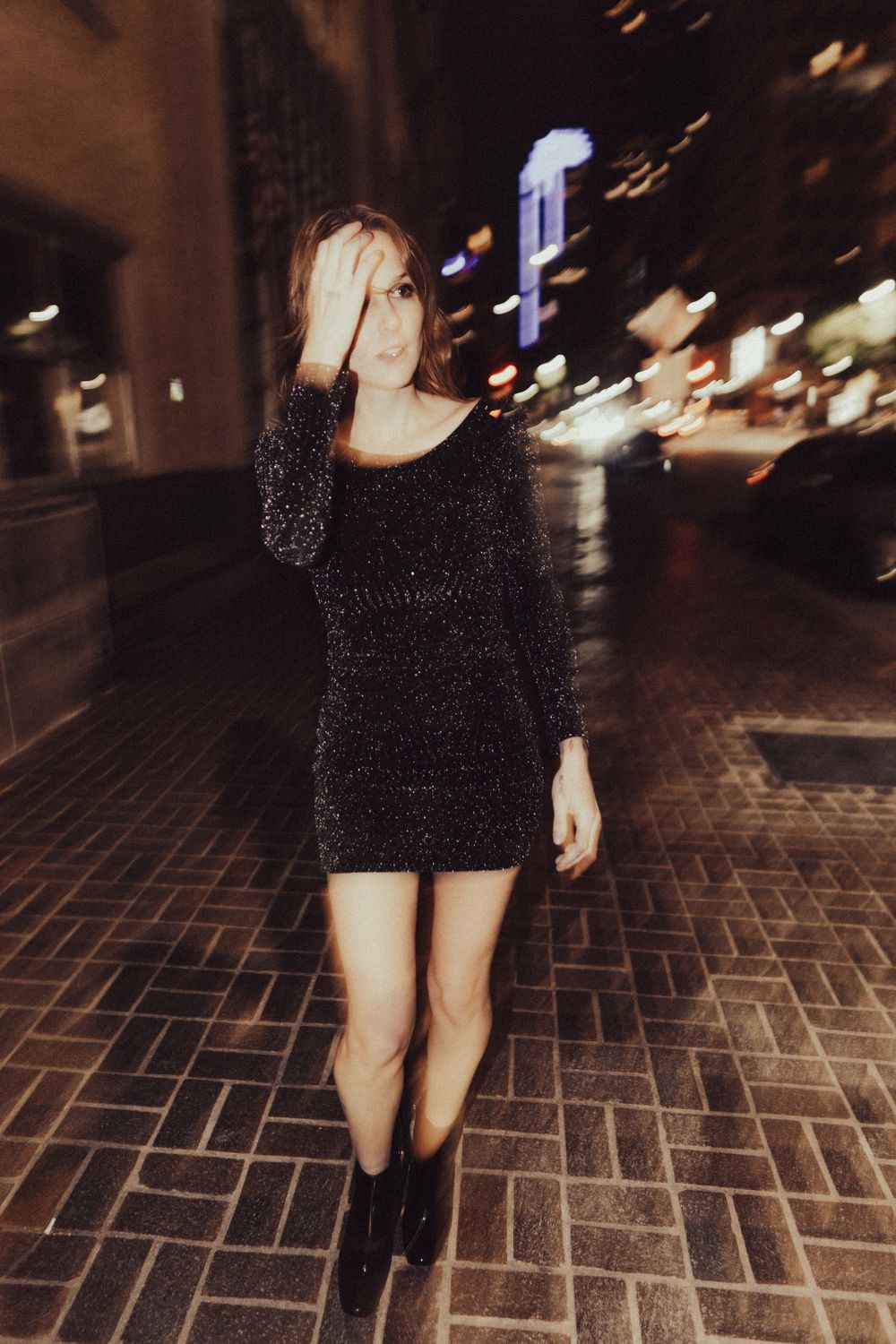 A figure in a glittery black mini dress poses on urban brick pavement at night.