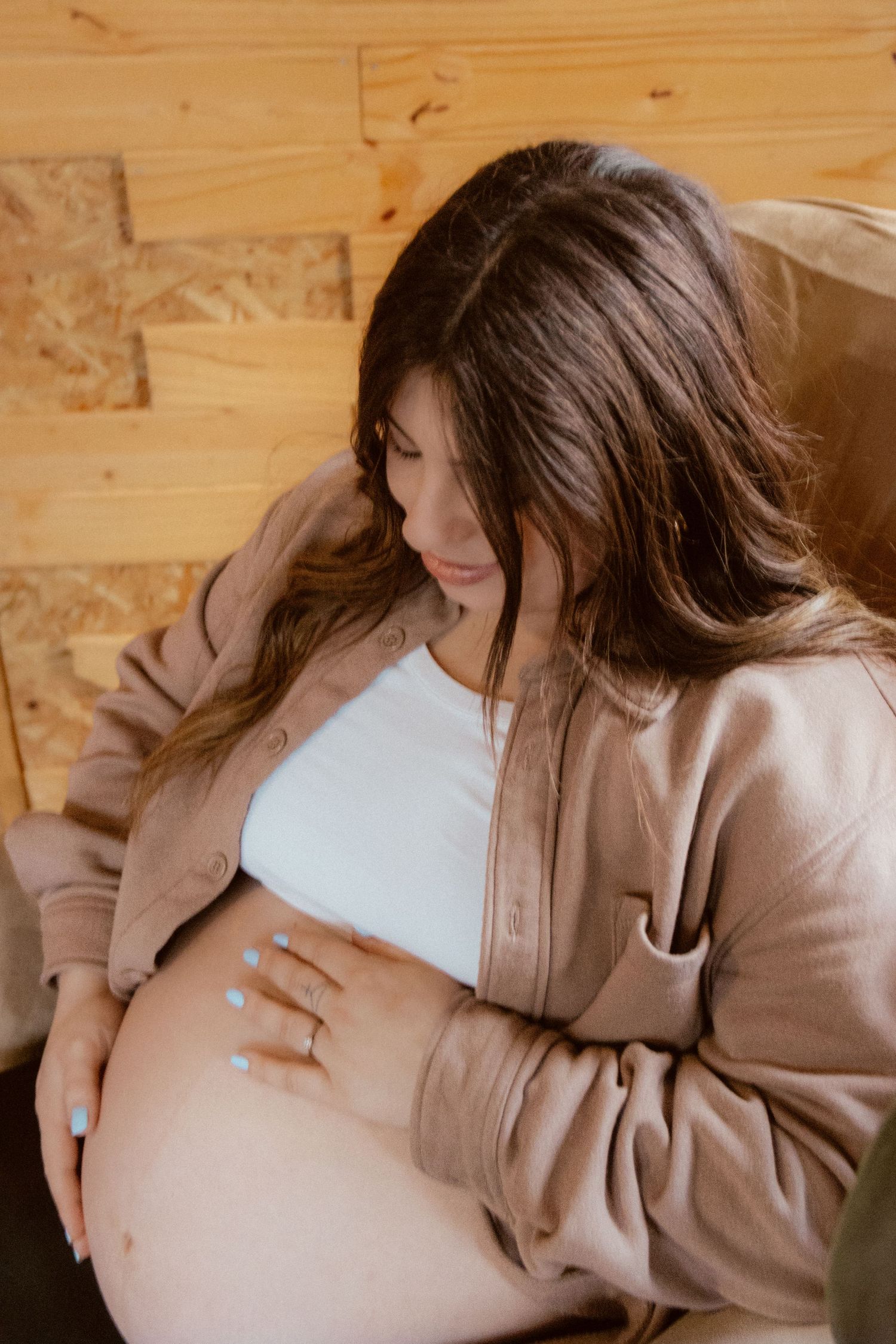 An expectant mother in a beige sweater gently touches her pregnant belly in warm indoor lighting.