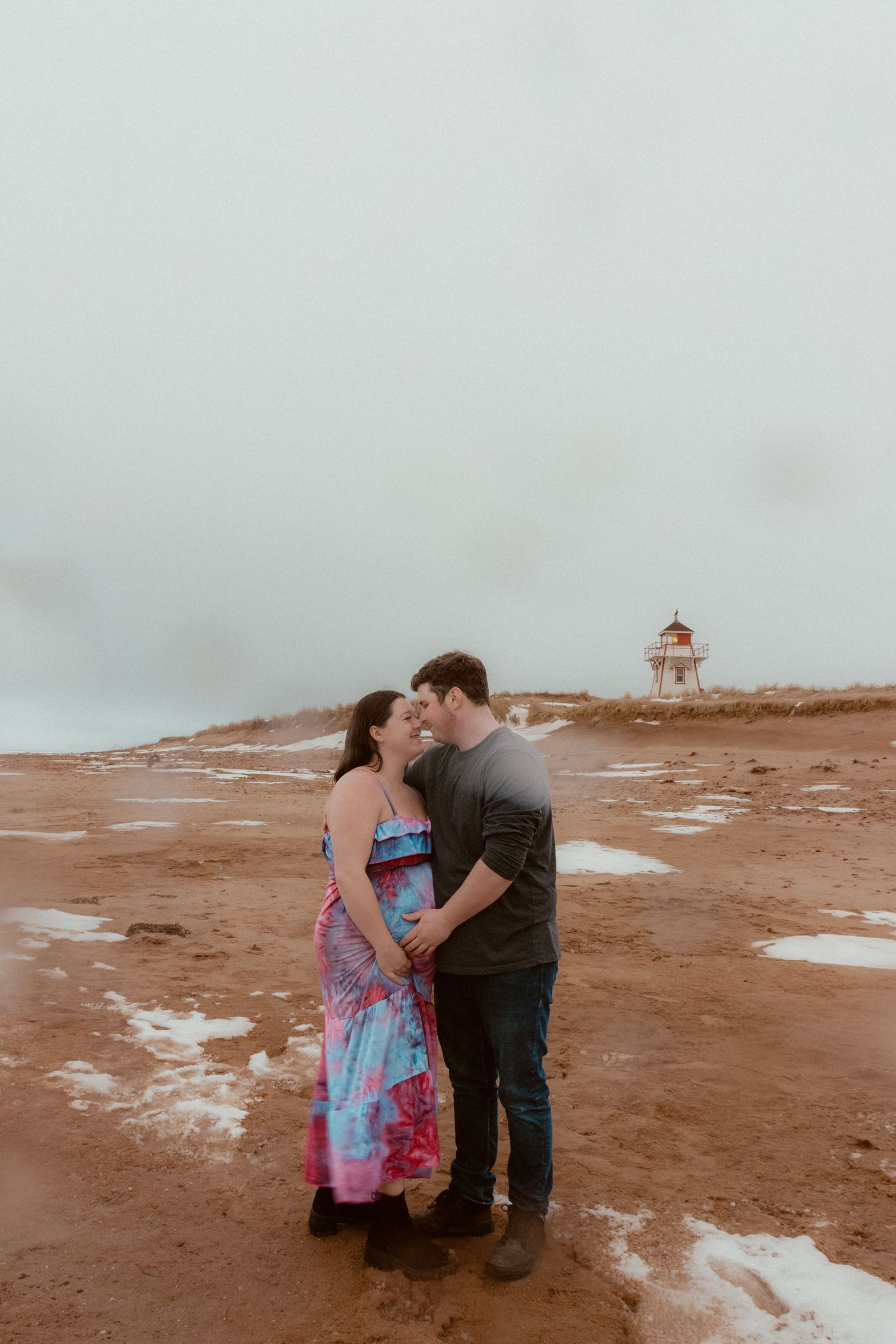 A couple embraces in front of a lighthouse on a cloudy day at the beach.