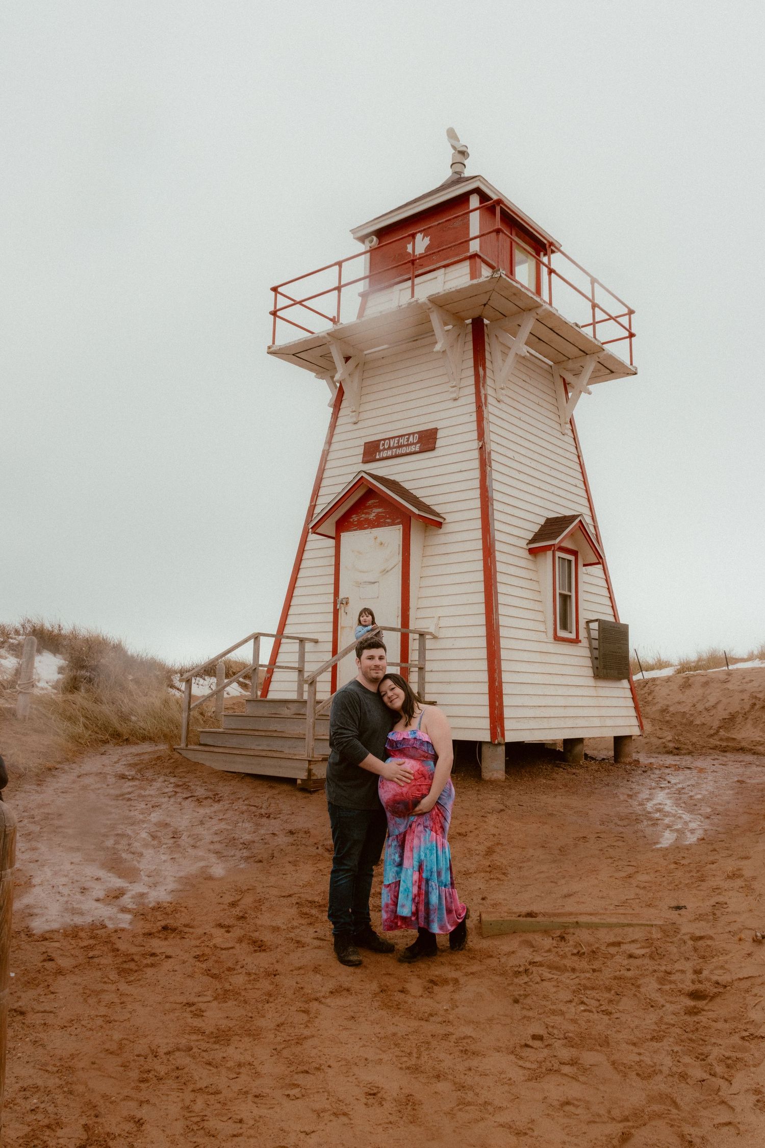 A couple poses for a photograph in front of a white lighthouse on a cloudy day.