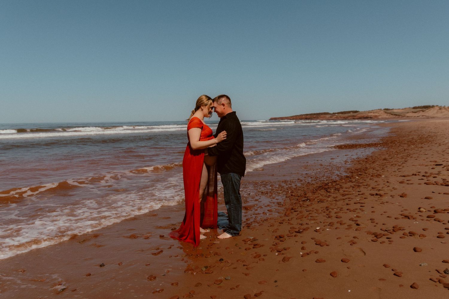 A couple in formal attire embraces on a beach with waves in the background.
