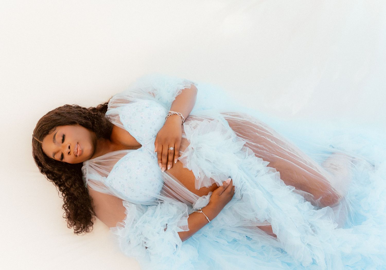 A graceful figure in an ethereal light blue tulle gown reclines against a white backdrop in a dreamy portrait setting.